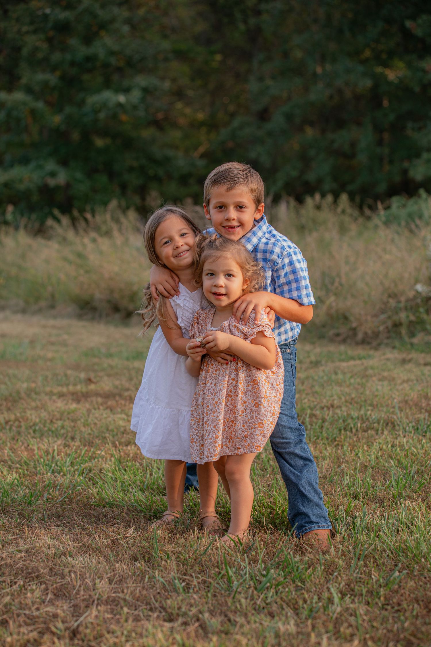 Three young children embrace in a sweet moment while standing in a grassy field at sunset.