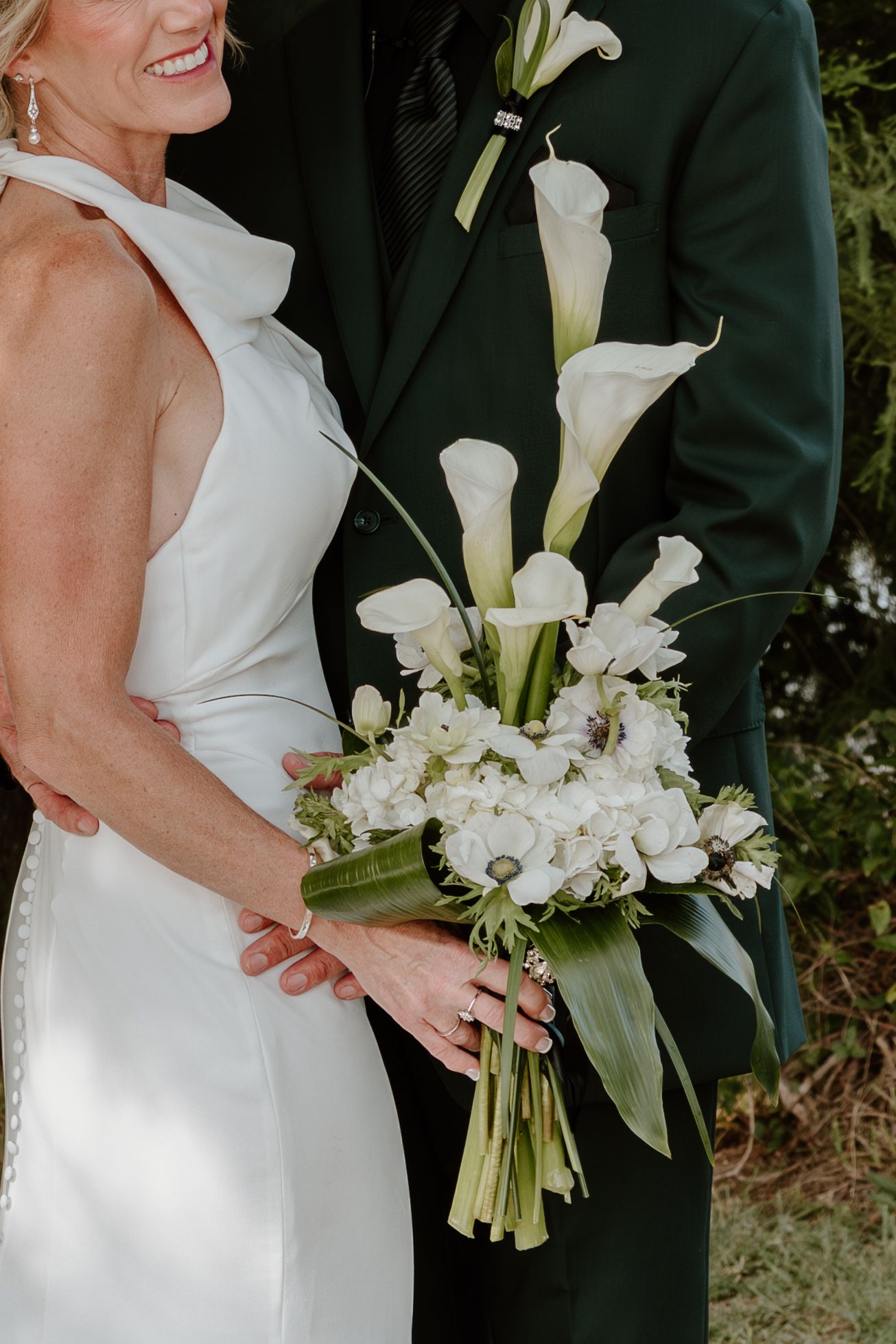 Bride holding a white calla lily and hydrangea wedding bouquet while standing with groom in dark suit.