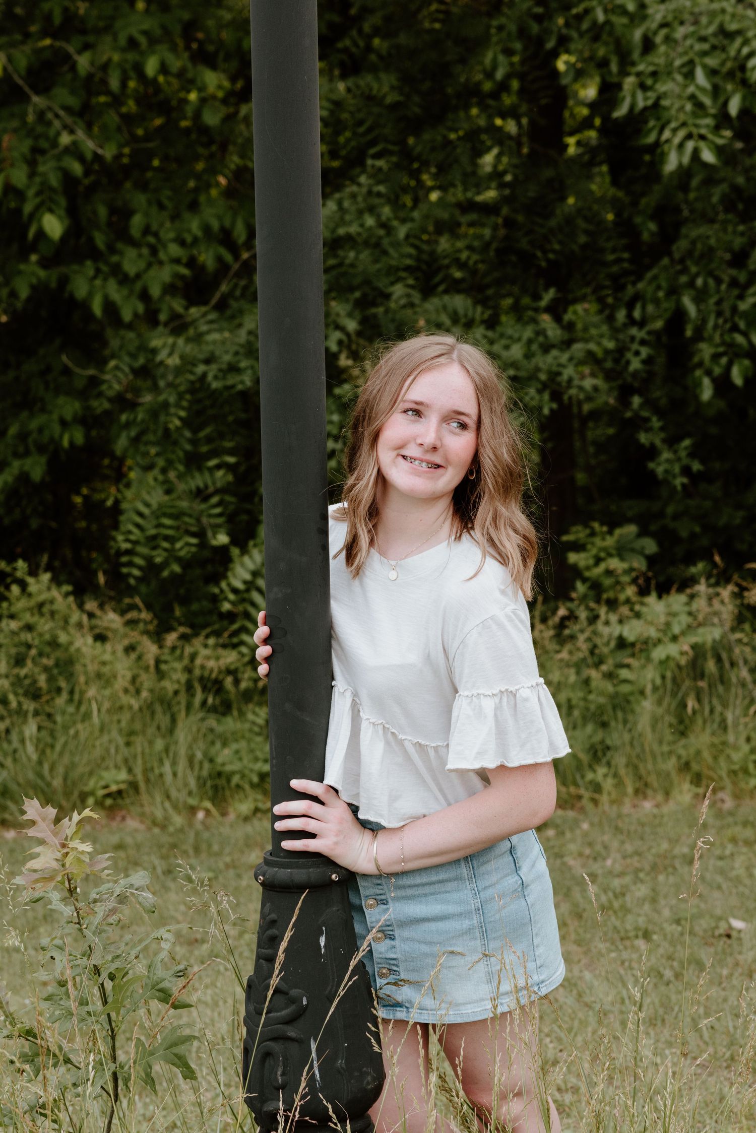 A child in a white blouse and denim skirt stands in a grassy field near a post surrounded by green foliage.