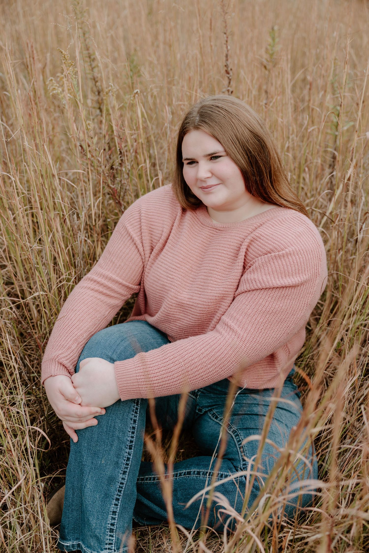 Someone wearing a pink sweater and blue jeans sits in tall dried grass during an outdoor photo session.