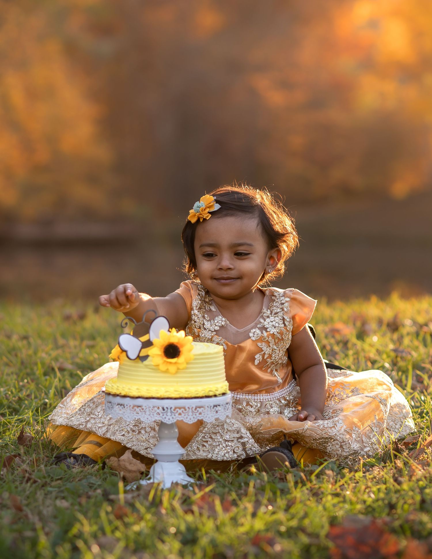 a little girl in a gold dress sits in front of a yellow smash cake with beautiful fall foliage in the background