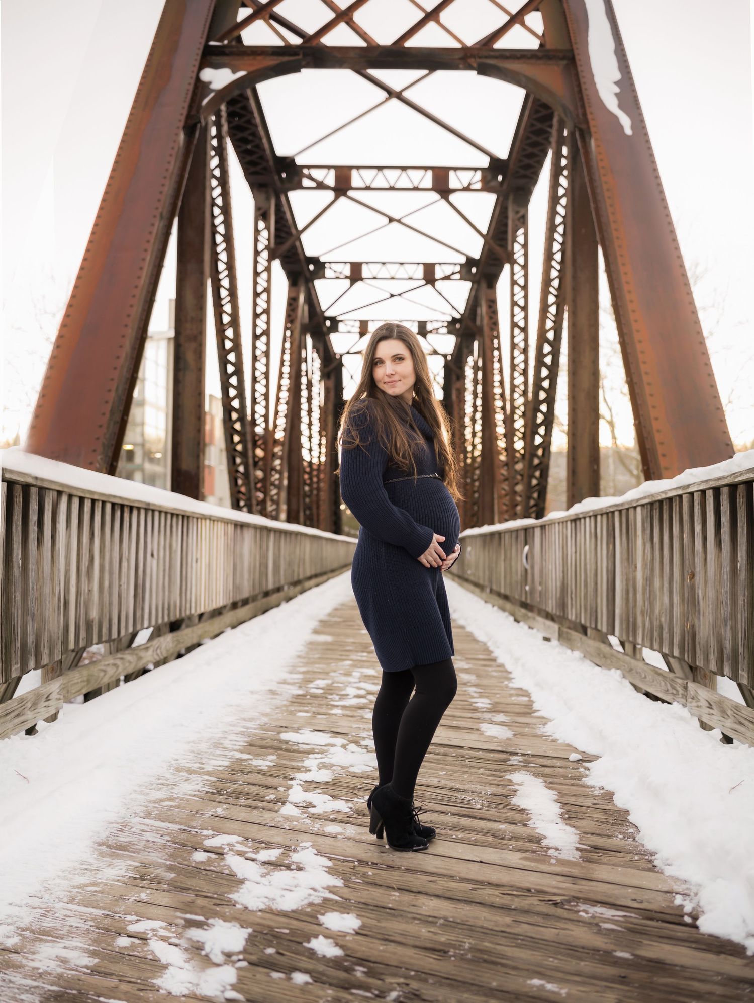a young pregnant woman with long dark hair poses for a maternity portrait on an old iron bridge with snow all around