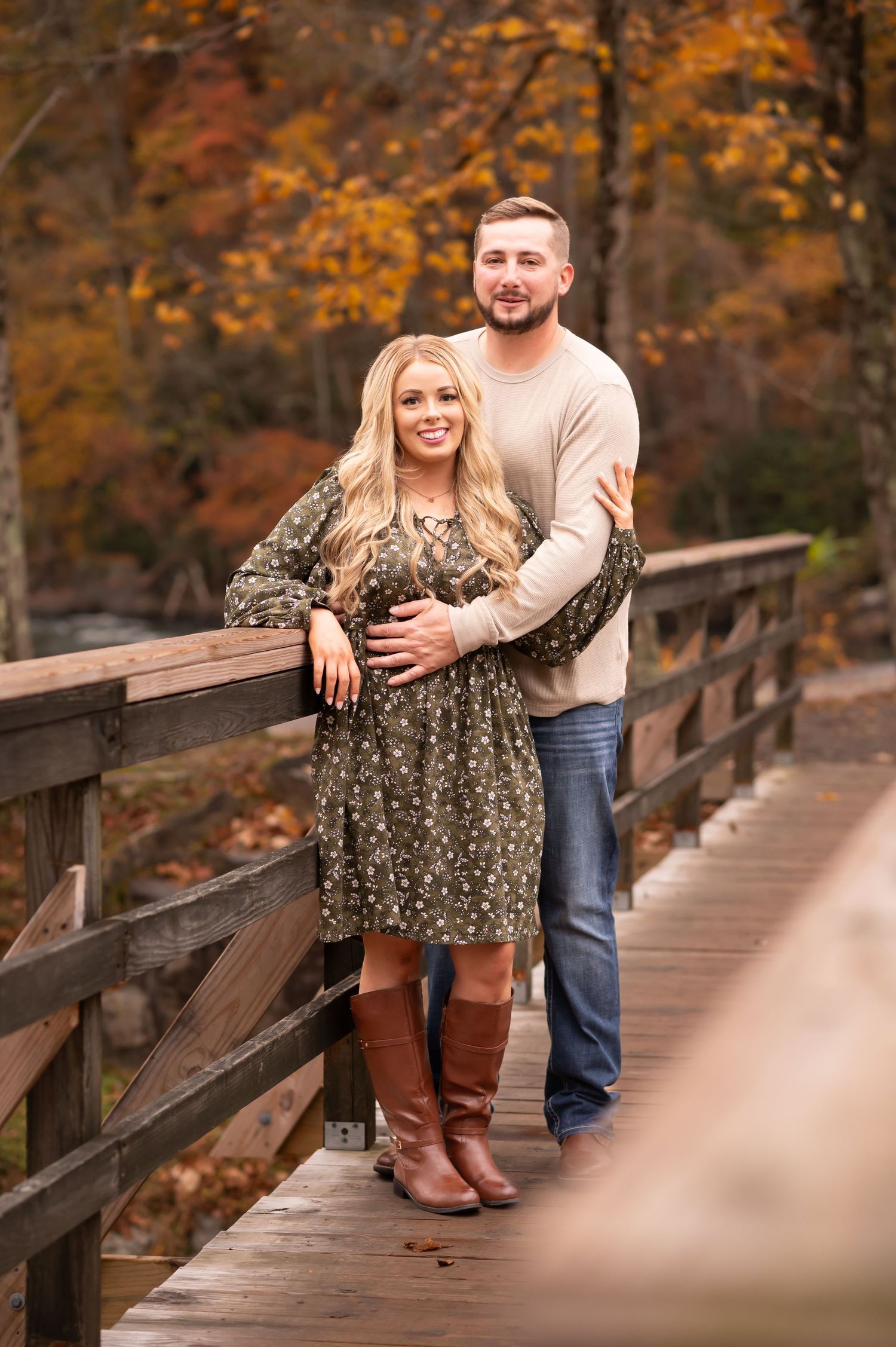 a young couple poses and smiles for a portrait together on a bridge with fall scenery behind them  in Valley Falls State Park