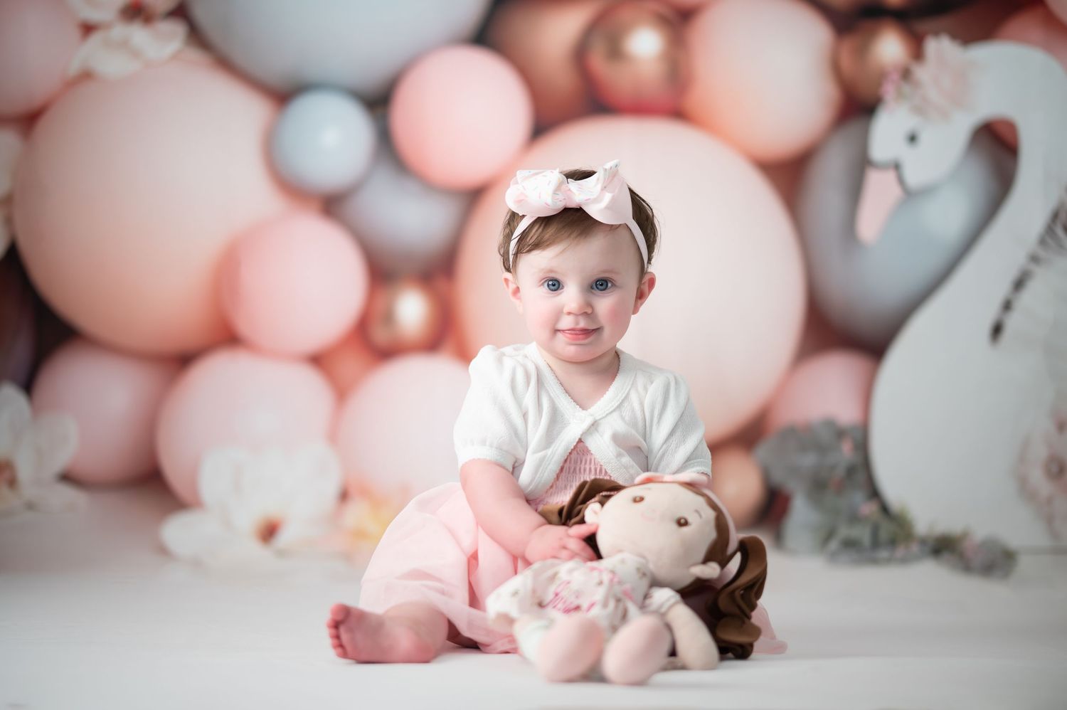 a little girl in a pink and white dress poses with her doll for a studio first birthday portrait with pastel balloons in the background