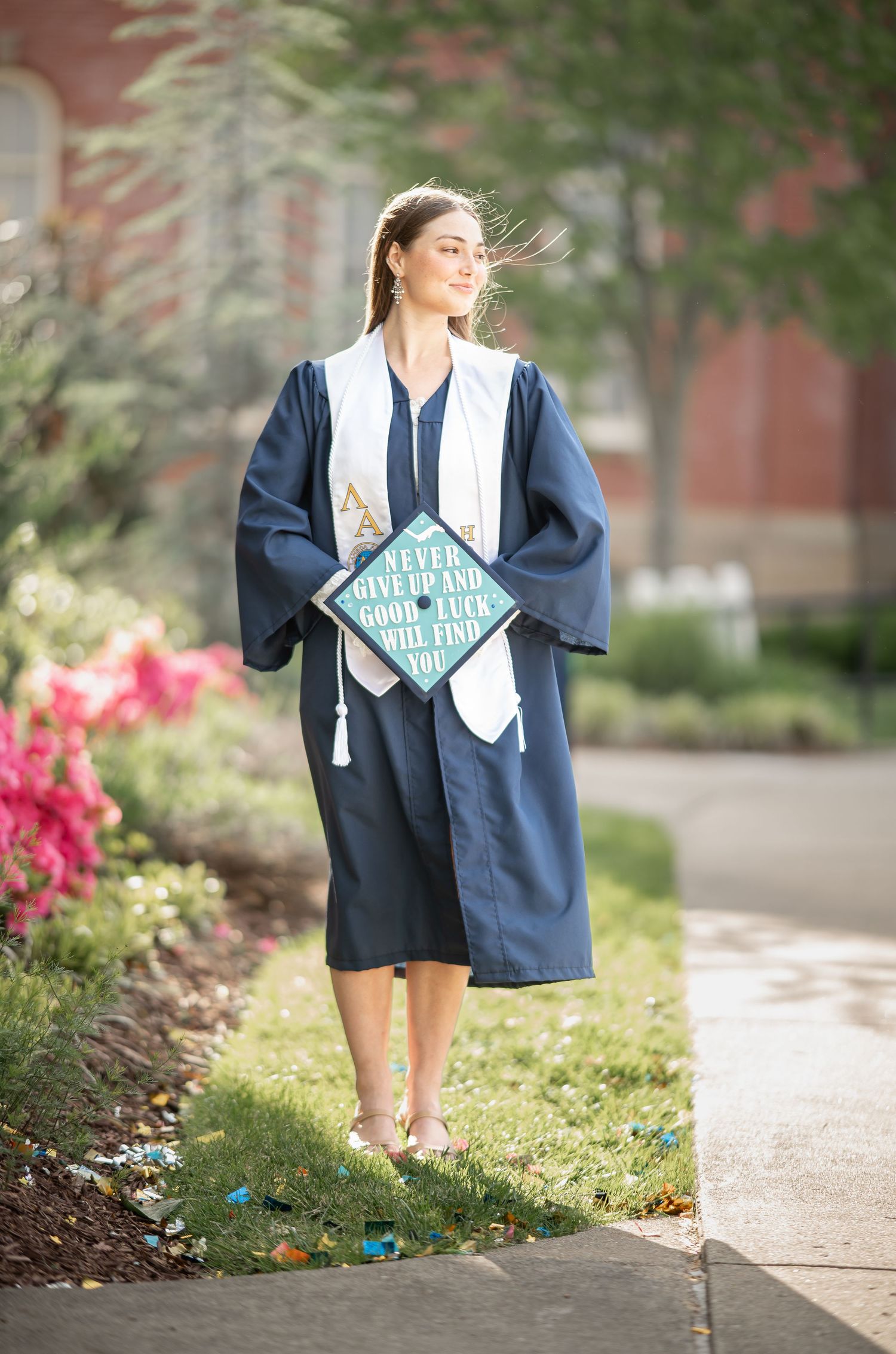 a WVU graduate student in her graduate gown stands beside bright pink flowers in the sunshine, holding her graduation cap