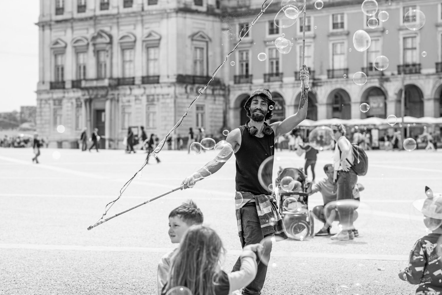 Street performer blowing giant bubbles in a busy plaza, with a child watching, captured in black and white.