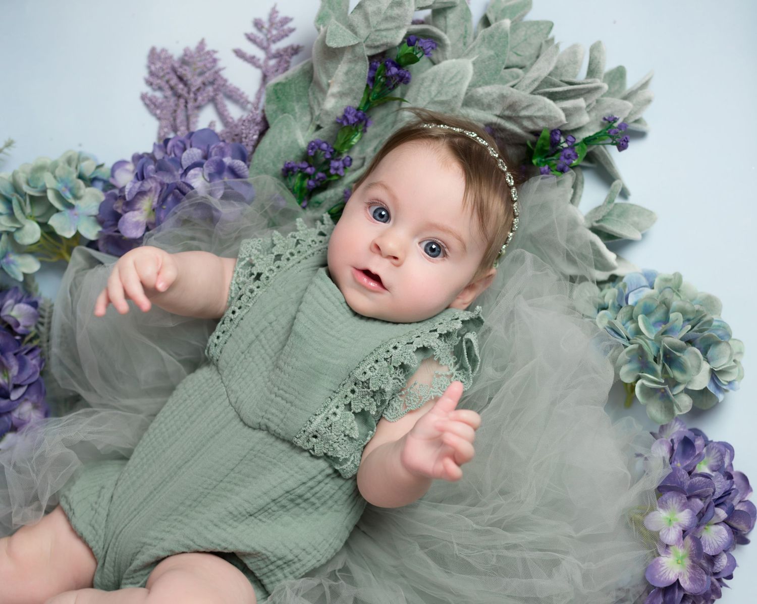 a baby girl in a green romper lies on a green blanket surrounded by purple and green flowers and looks up at the camera