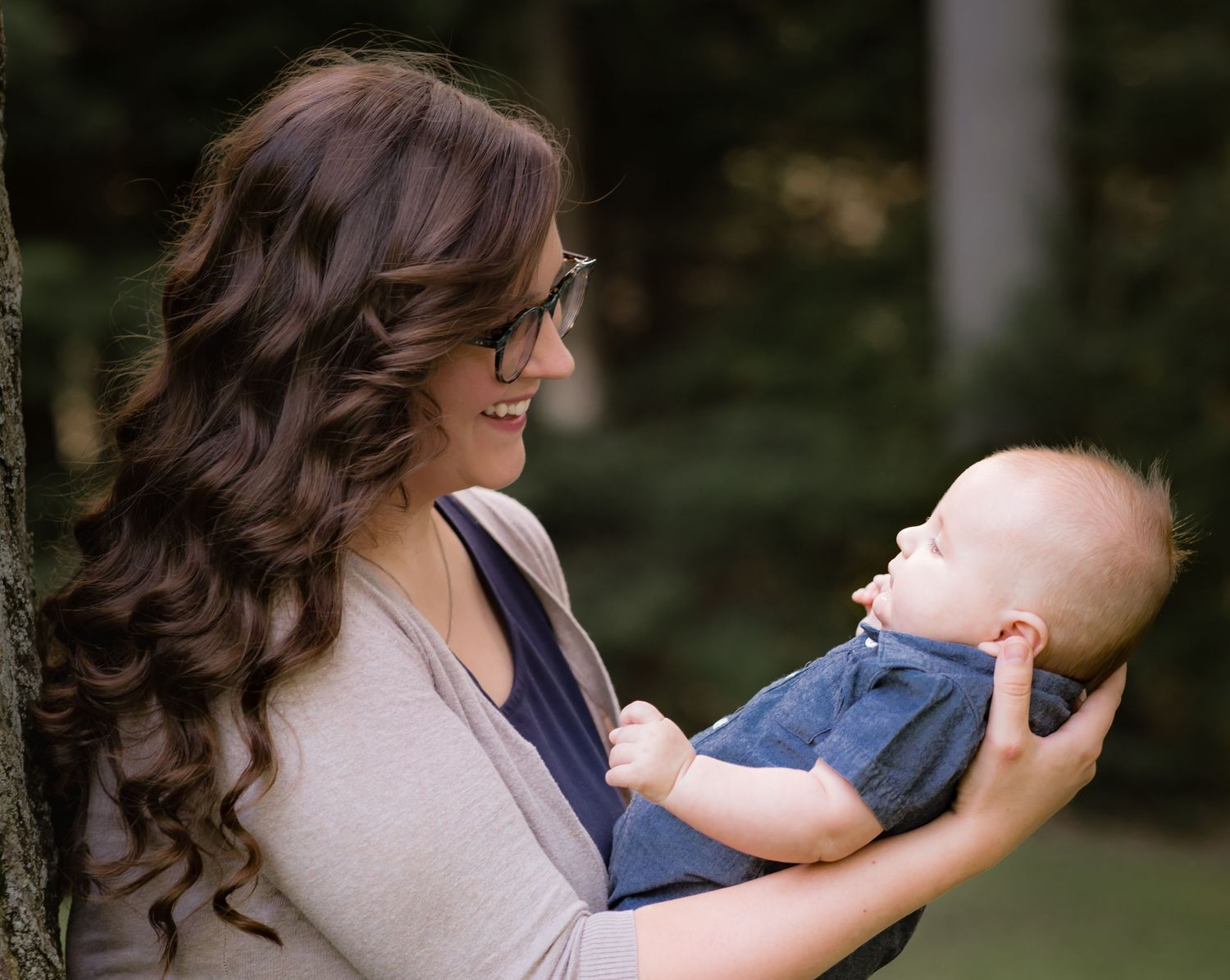a mother with long dark hair holds her baby boy and smiles at him, with green woods in the background