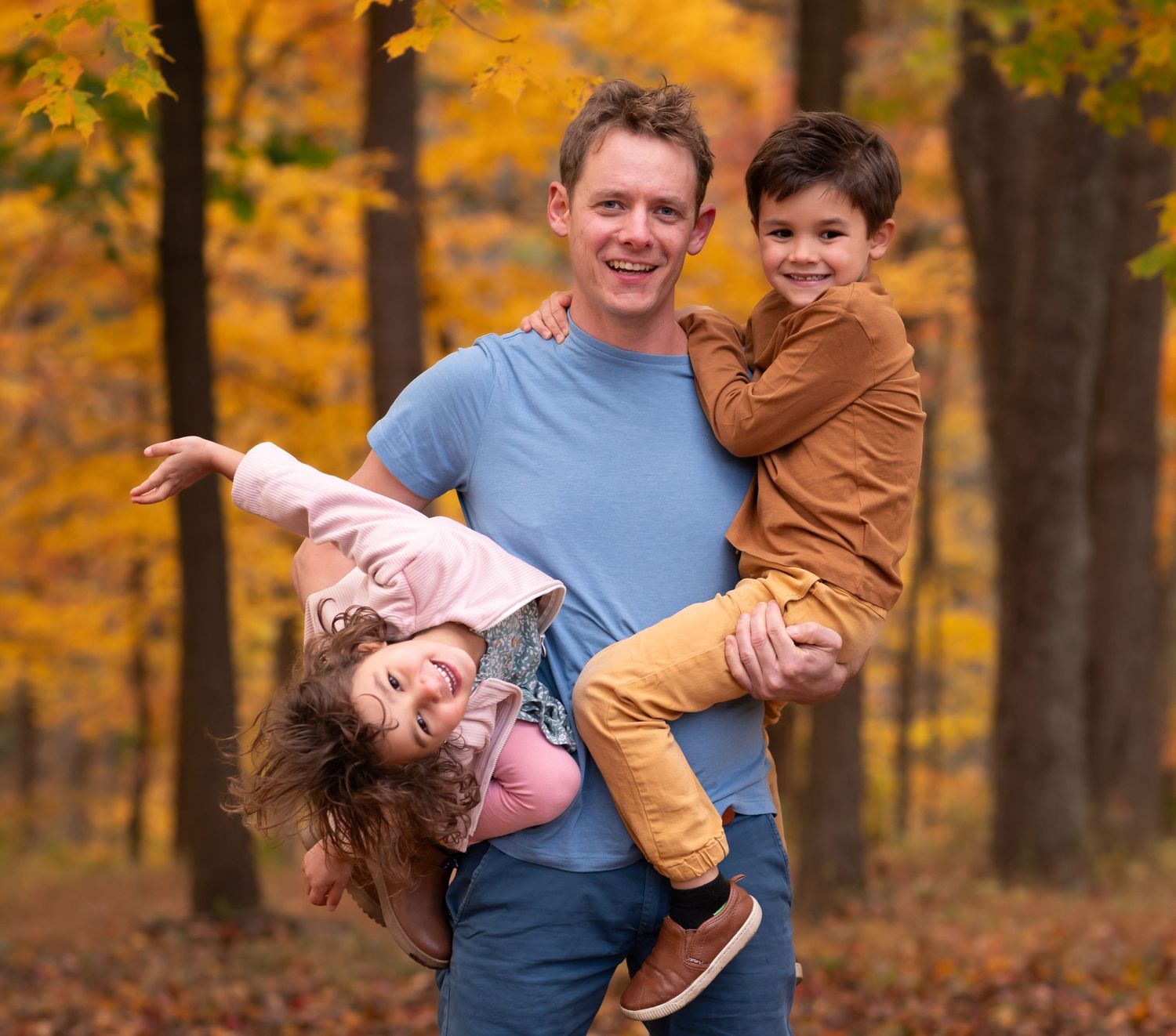 candid portrait of a young father holding his two children as they laugh and goof off, with fall scenery in the background