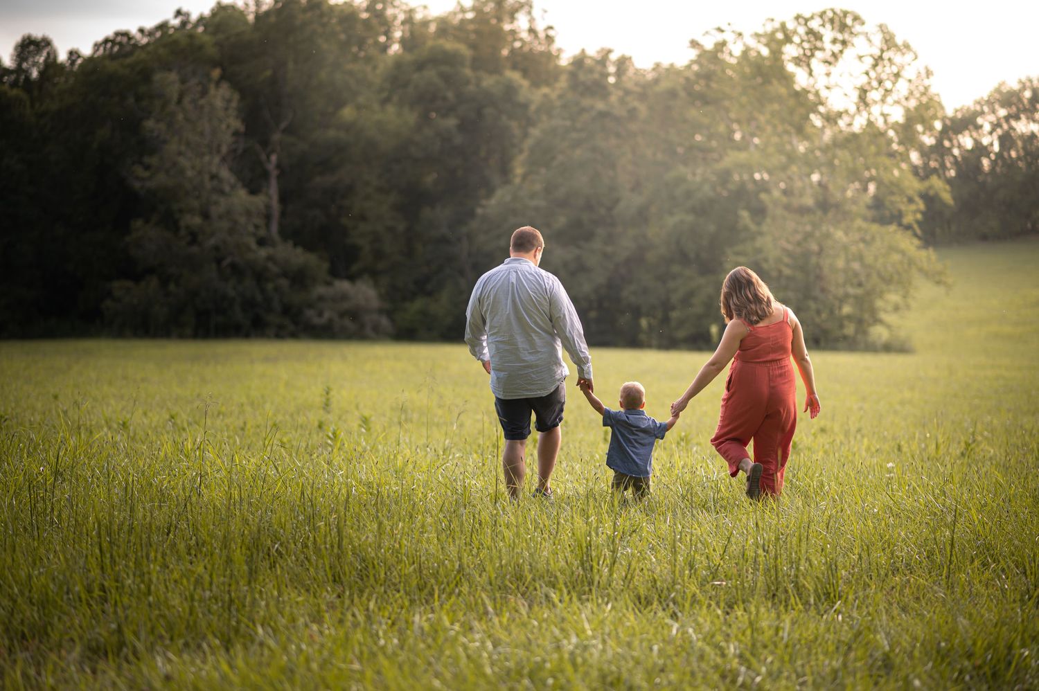 two parents hold their little son's hands and walk away through a field of tall green grass as the sun sets in front of them.