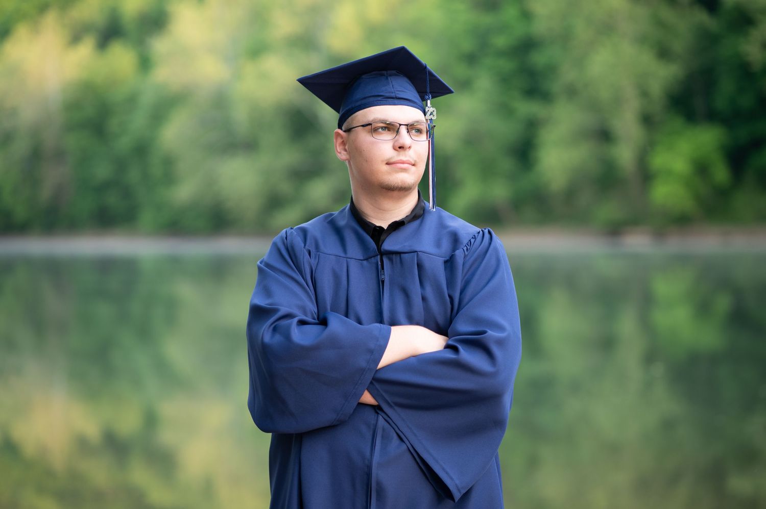 homeschool high school graduate in a navy cap and gown poses with arms crossed in front of a placid river with trees reflected in the water