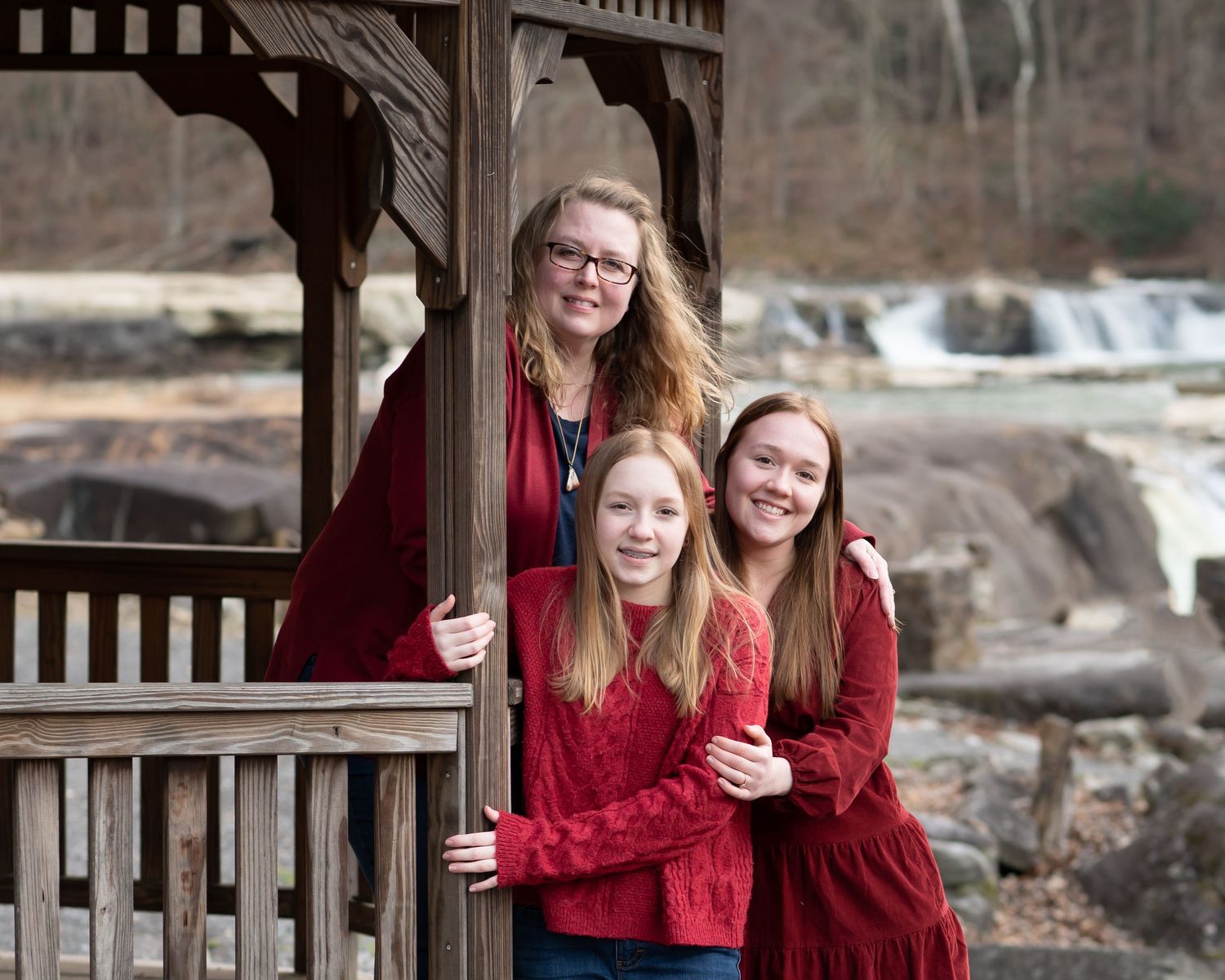 a mother and her two teenage daughters in red sweaters pose together for a family portrait in an old gazebo with a river in the background