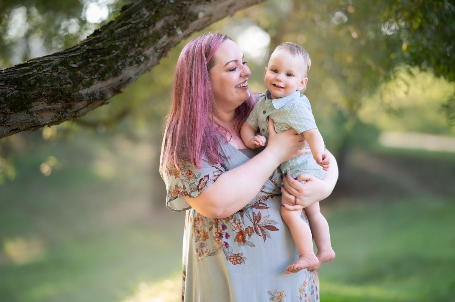 a young mother in a gray dress stands under a tree holding her toddler son as he smiles at the camera; shot in Morgantown, WV