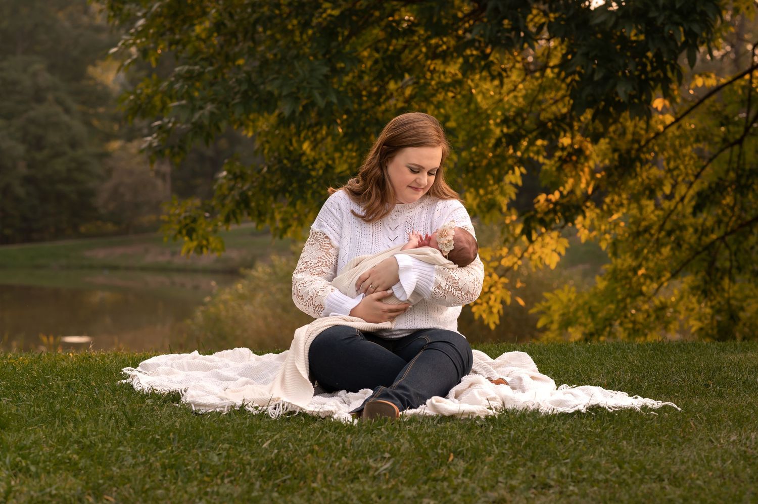 a young mother sits with her newborn baby girl on a white blanket in the grass, with fall foliage behind her, in Morgantown WV