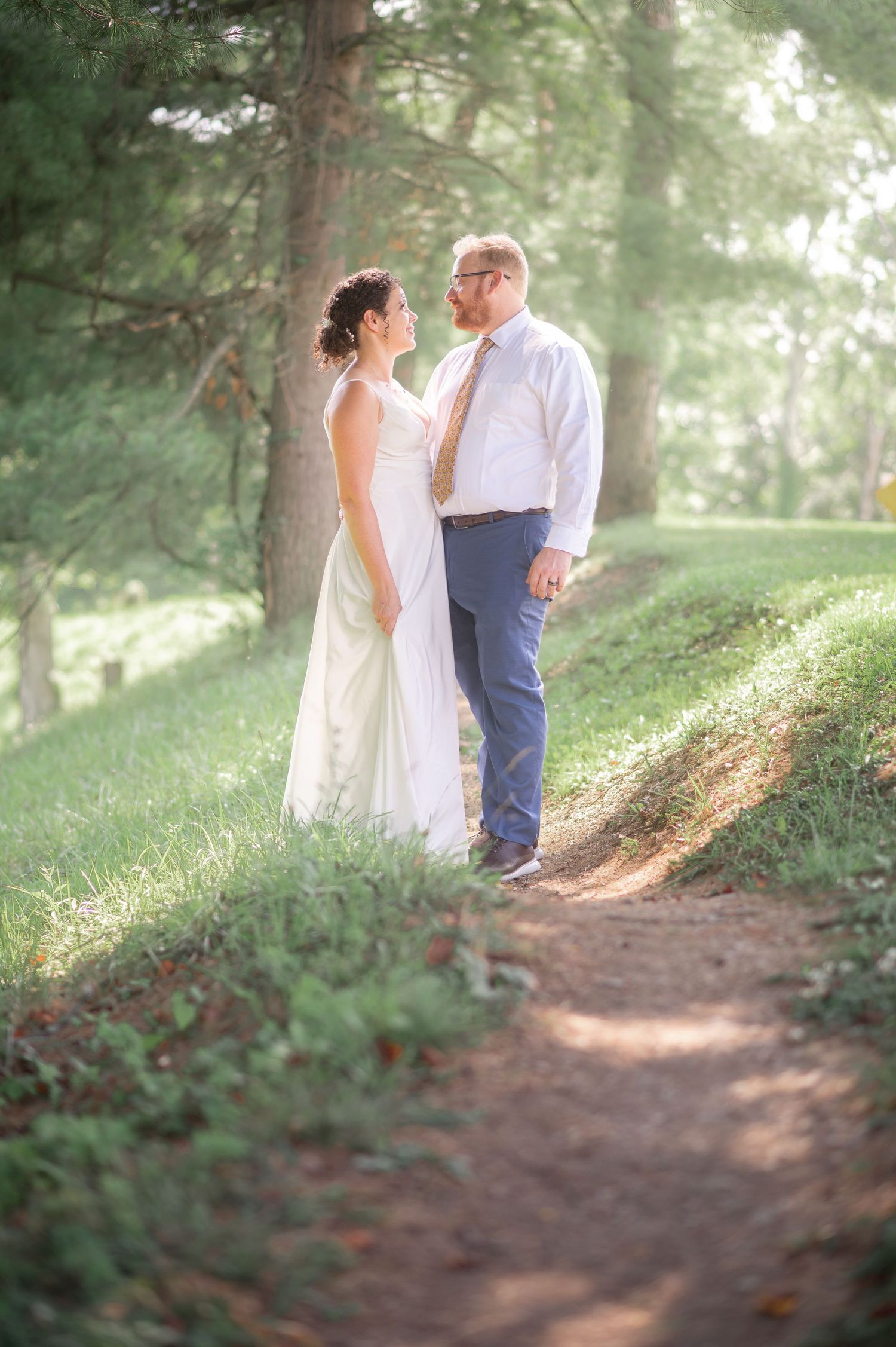 a bride and groom stand on a wooded path awash with sunlight, with misty trees behind them, and look at one another