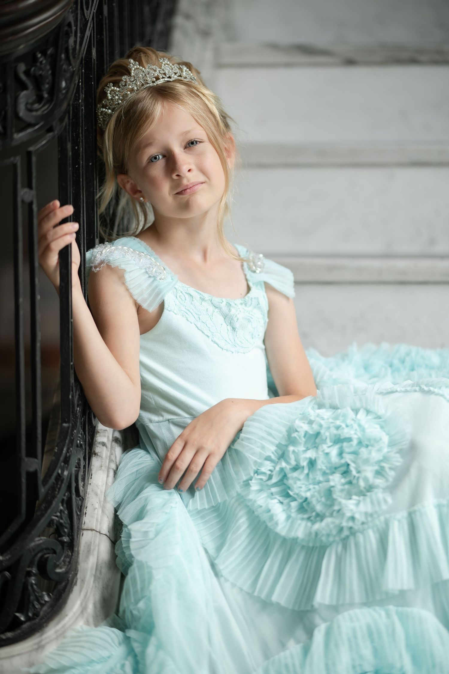 A young girl in a light blue princess gown and tiara sits gracefully beside a dark ornate post.