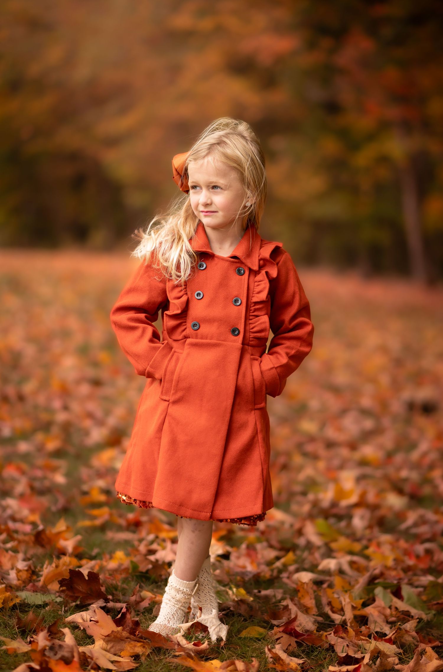 product shot of an orange pea coat worn by a little blonde girl in white tights and boots, standing surrounded by autumn leaves and trees