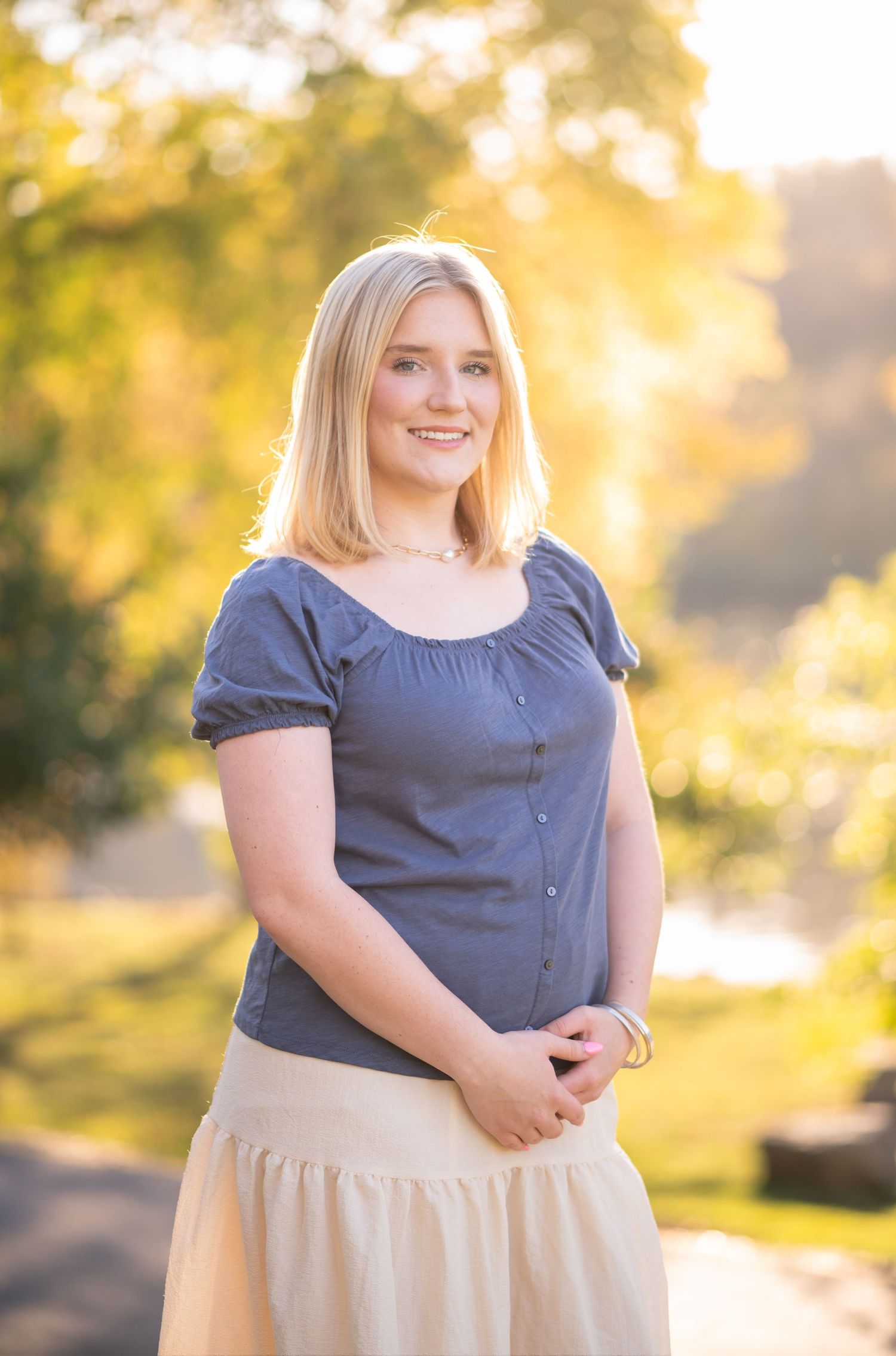 a young blonde woman in a blue top and white skirt poses and smiles for a portrait with sunlit golden leaves in the background
