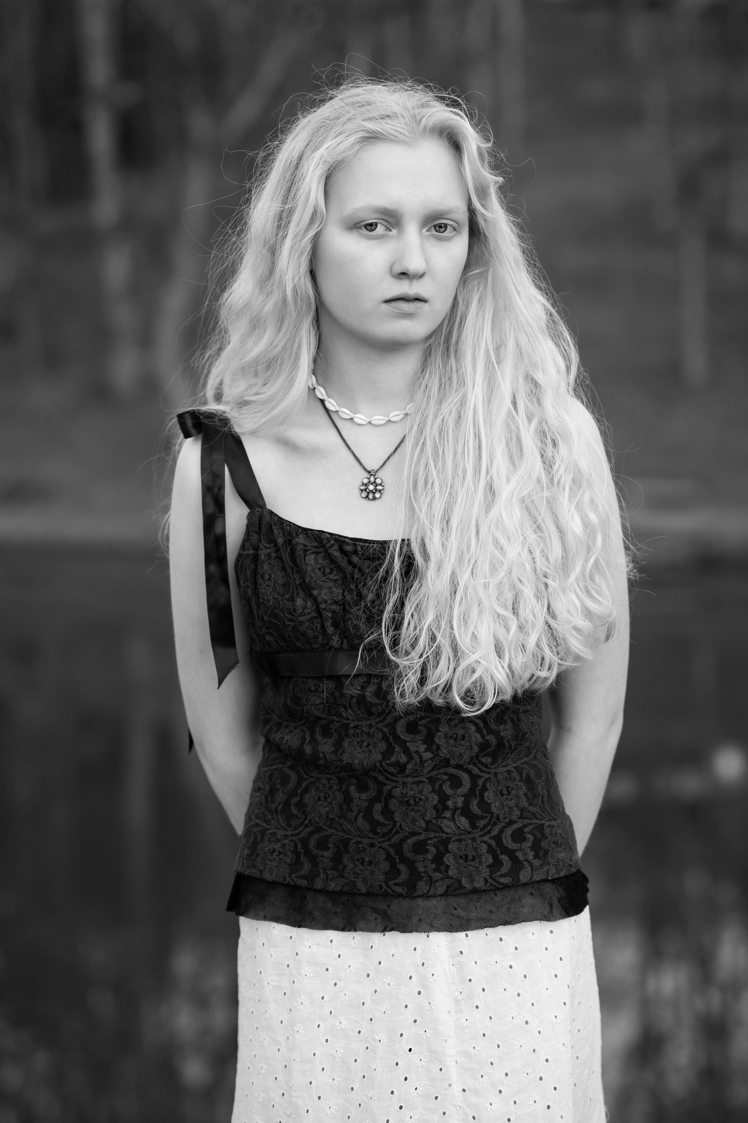 thoughtful black and white portrait of a young blonde woman in a black top, posing with hands behind her back and a pensive expression
