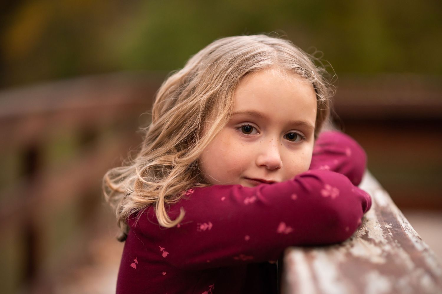 closeup portrait of a little girl, leaning her cheek on her arms against a wooden railing outside, with autumn foliage in the background