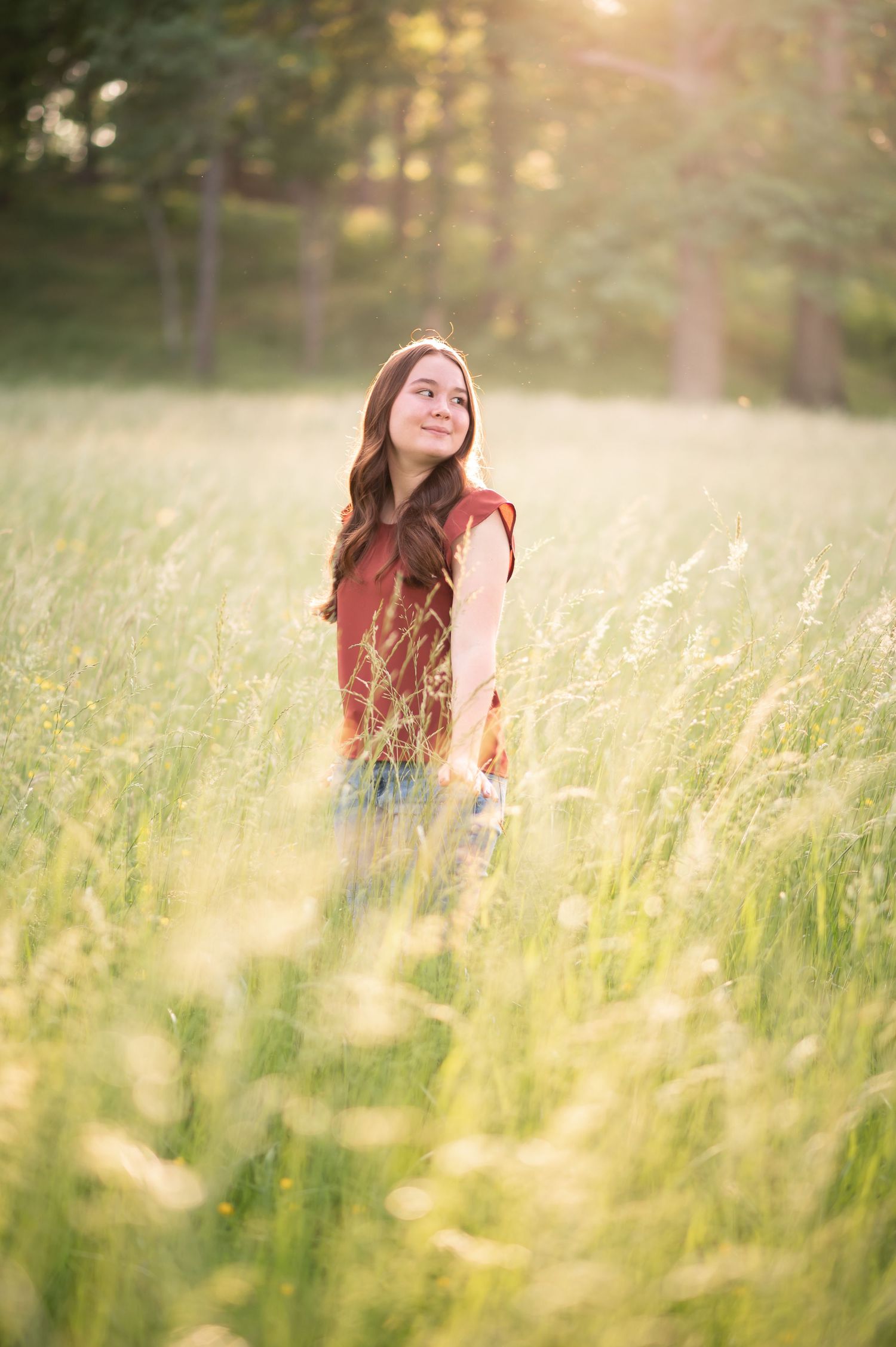 a lovely teenage girl in a ruffled orange top smiles in a sunlit field of tall golden grass, with forest in the background