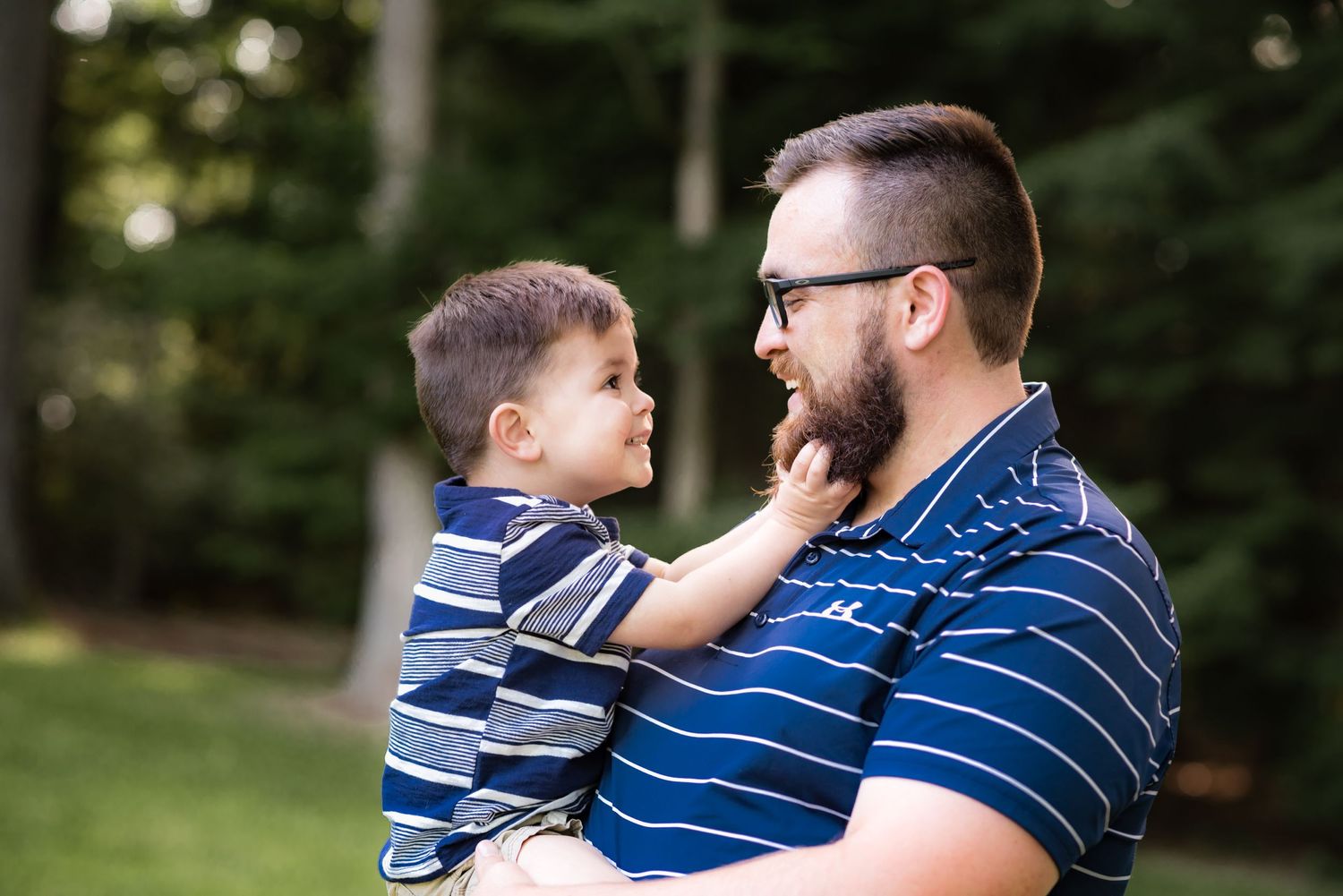 candid portrait pf a young father holding his little toddler son, while the son reaches up to grab his beard and they smile at each other.