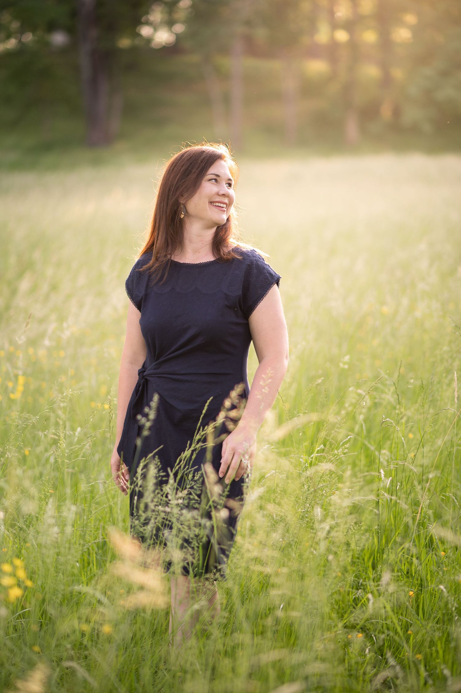 a lovely woman in a long navy sheath dress walks through a sunlit field of tall grass and laughs