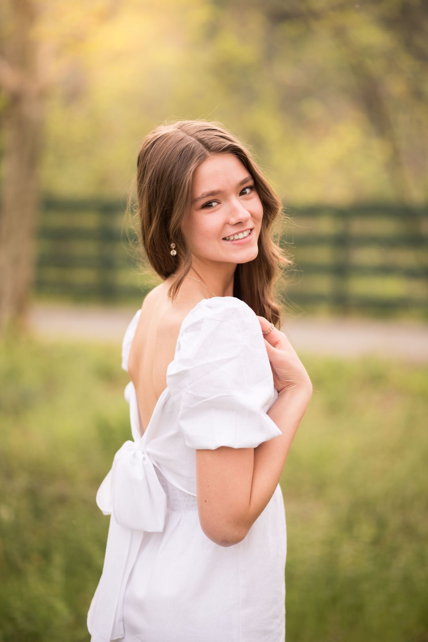 senior portrait of a young woman in a white dress, looking back over her shoulder and smiling at the camera, with a picket fence behind her