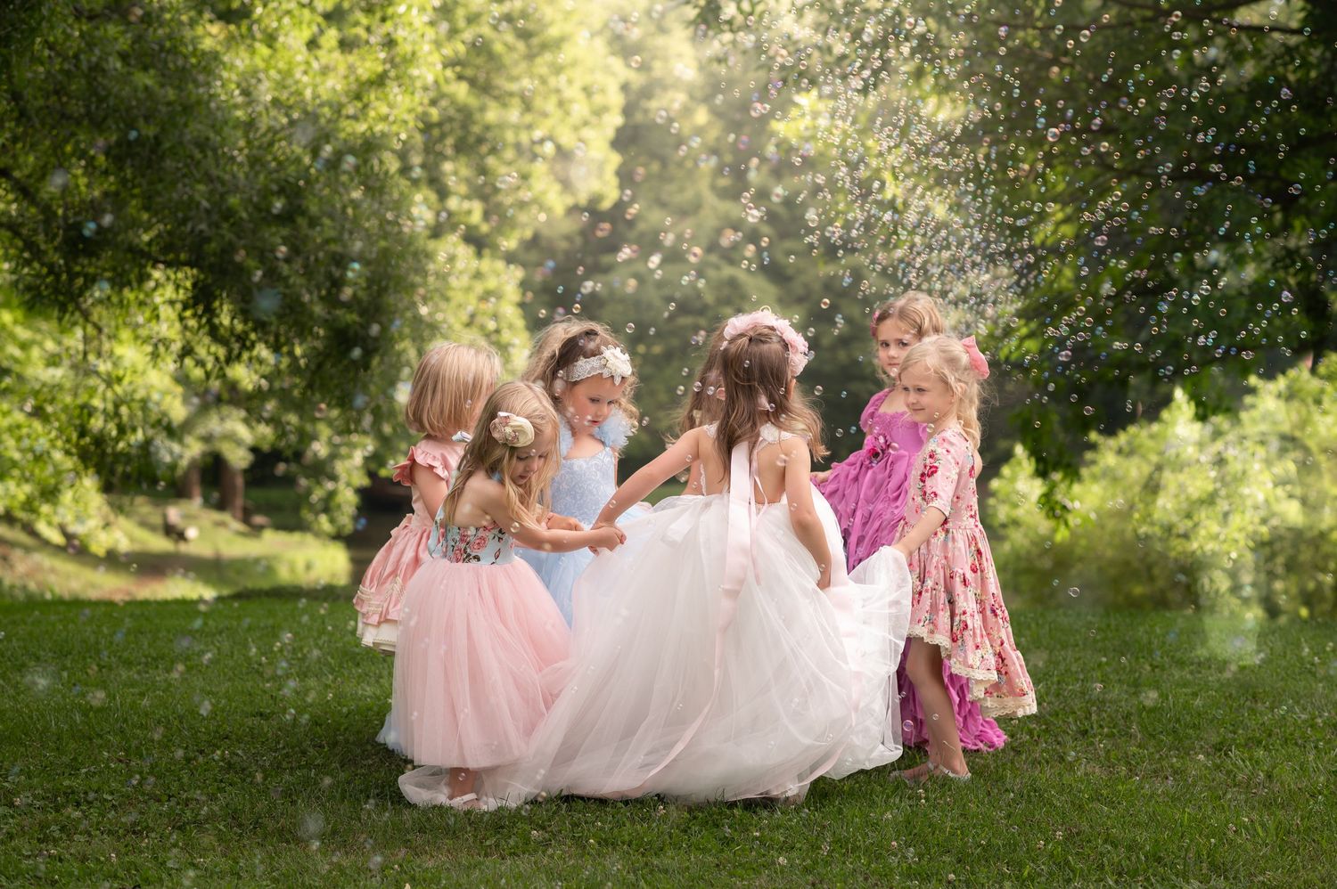 a group of little girls in fluffy floor length dresses hold hands and play ring around the rosie in lush green grass with a pond behind