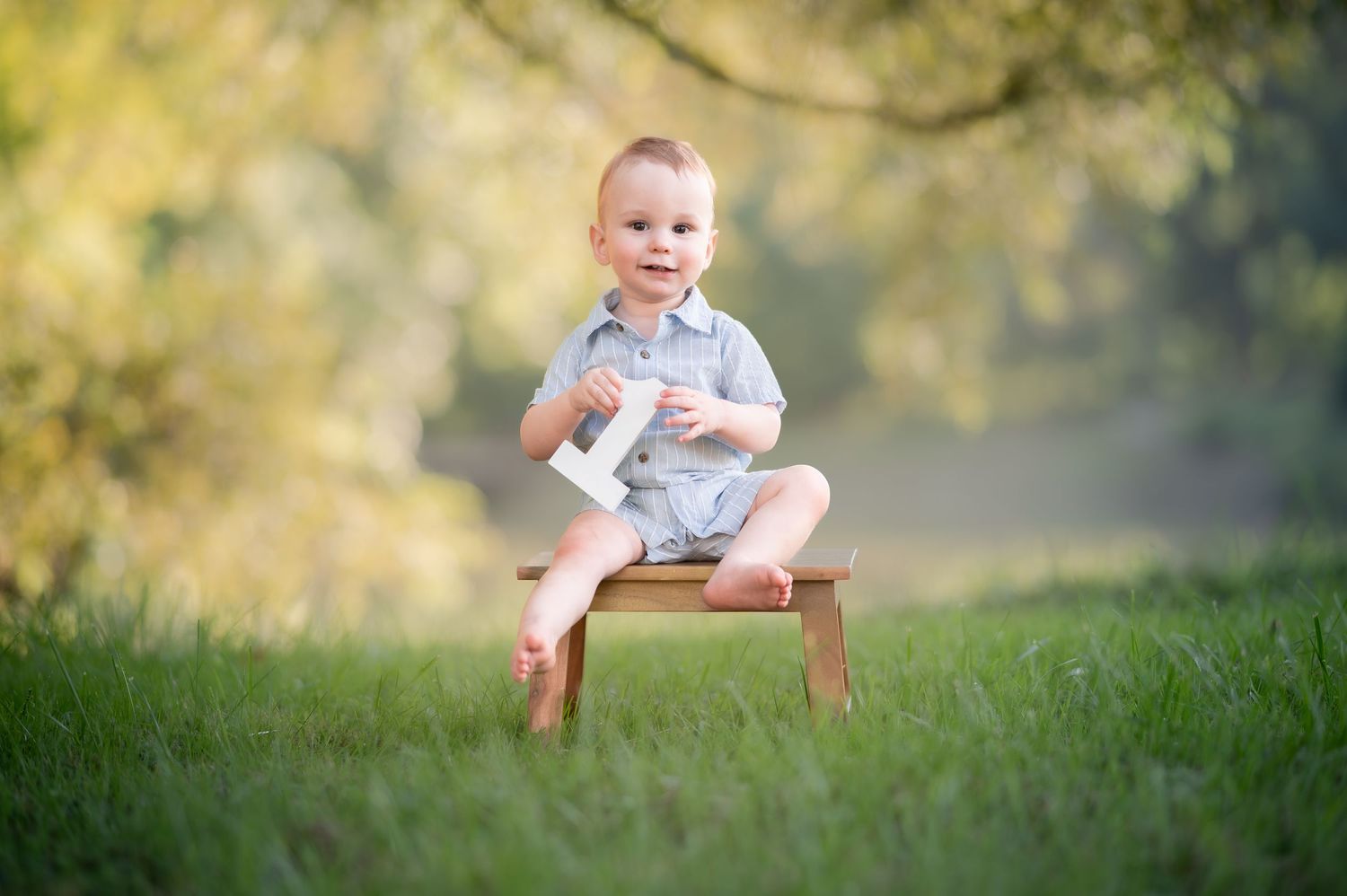 a one year old boy in a blue romper sits on a little wood bench holding a white wooden 