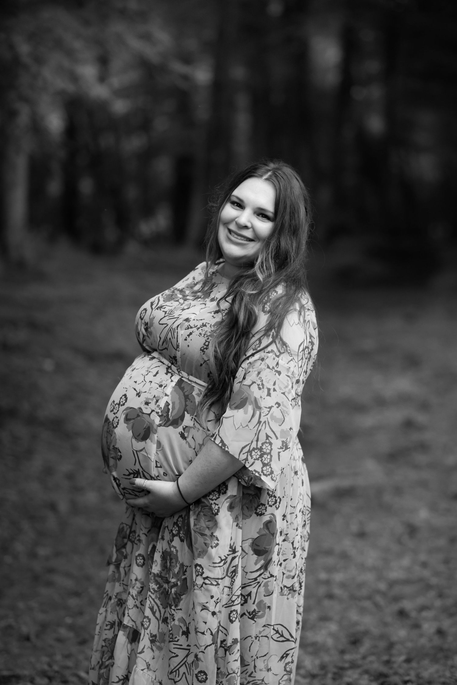 black and white maternity portrait of a woman in a long floral gown, posing with her hands on her belly