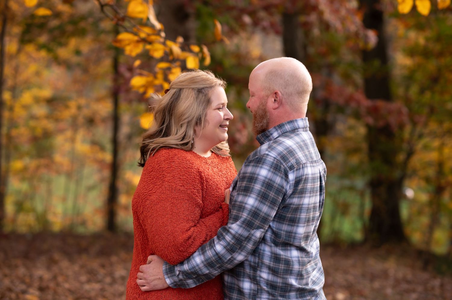 an engaged couple in fall sweaters stands together amid autumn foliage in a park and smiles at one another