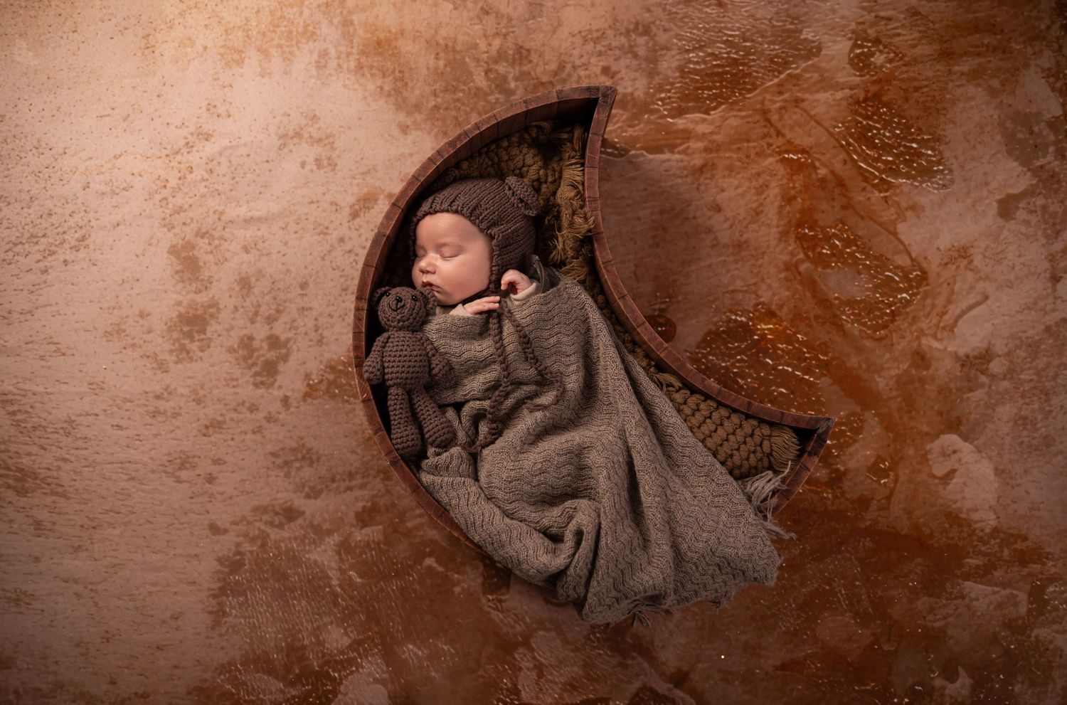 newborn studio portrait of a baby boy swaddled in brown, with a bear cap on his head, lying in a brown wooden moon in Morgantown, WV