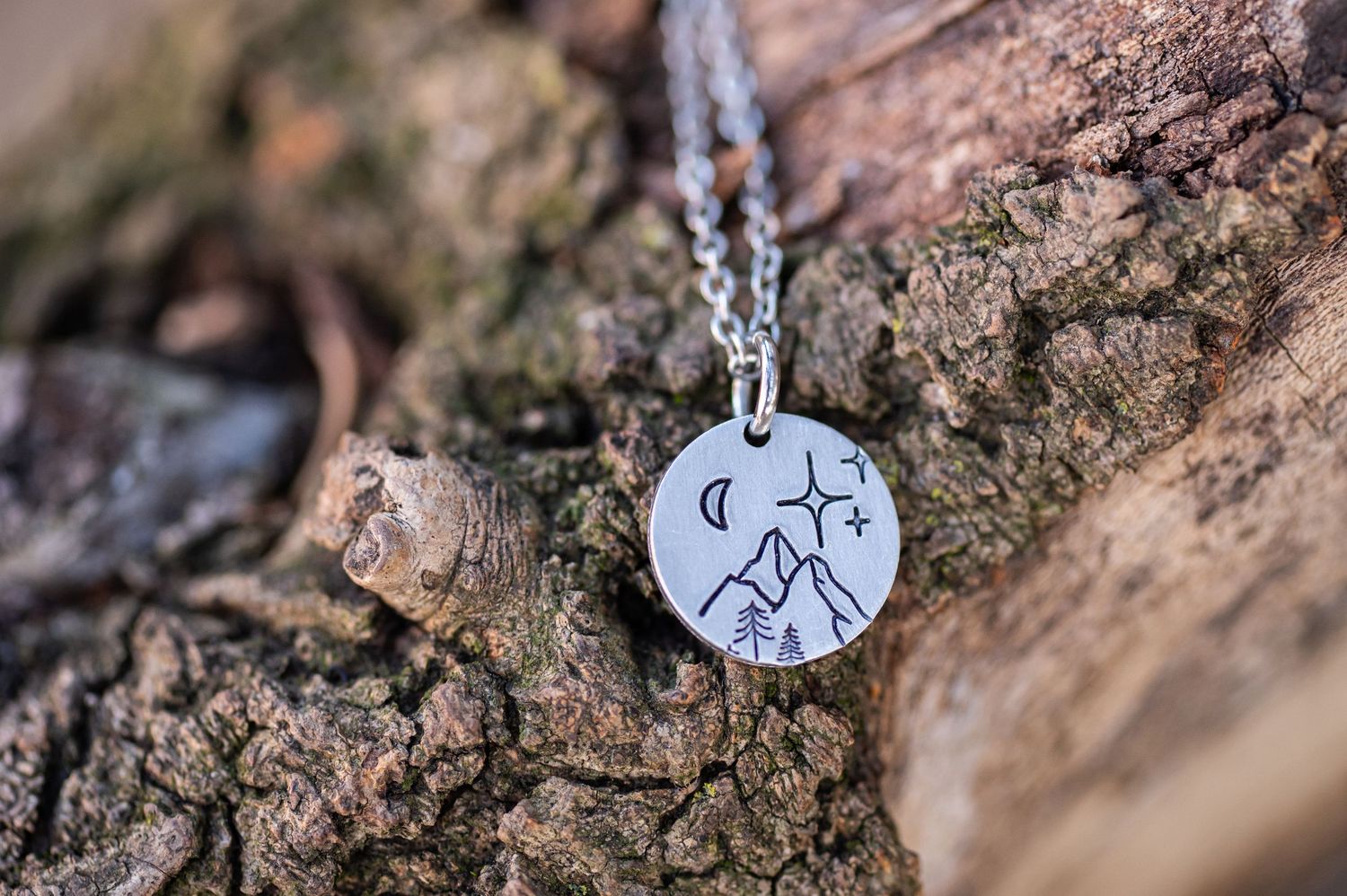 hand stamped silver necklace with mountains and stars lies on the rough bark of a tree branch