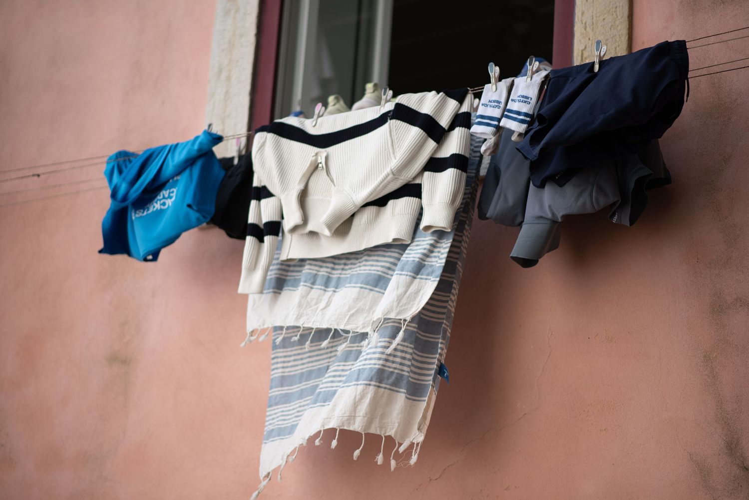 Colorful laundry including a striped blanket and blue clothes hanging on a clothesline outside a pink building window.