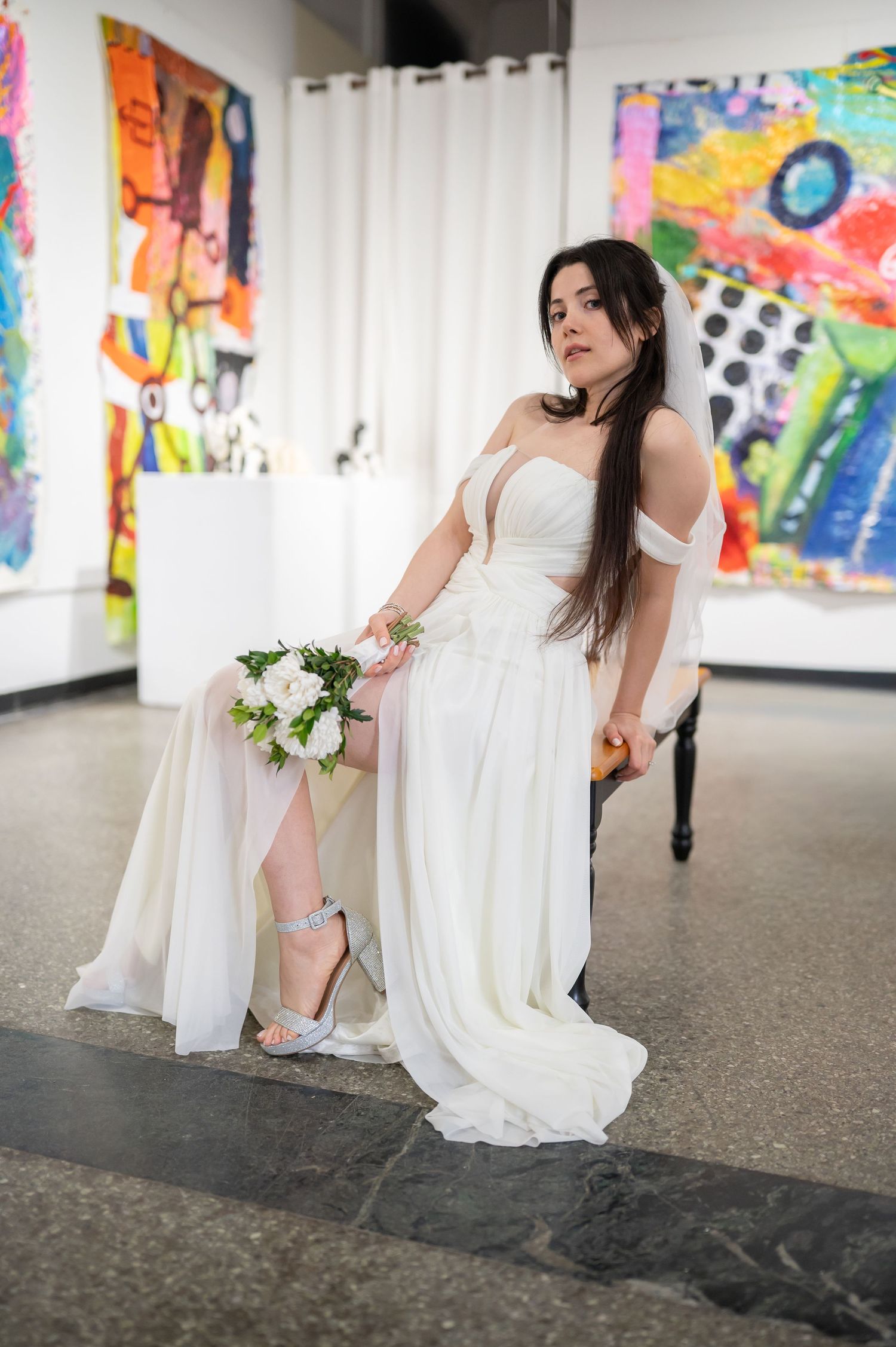 artistic portrait of a dark-haired young bride in a soft white wedding gown, posed on a stool in an art gallery and surrounded by bright art