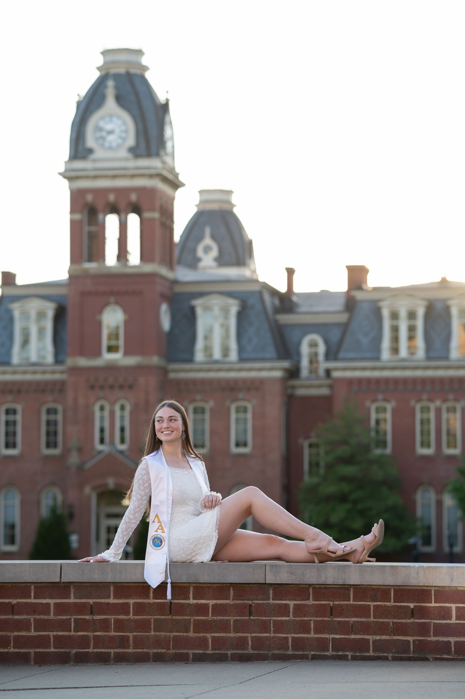 a graduate student in a white dress sits on a low brick wall with Woodburn Hall in the background; Morgantown, WV graduation sessions