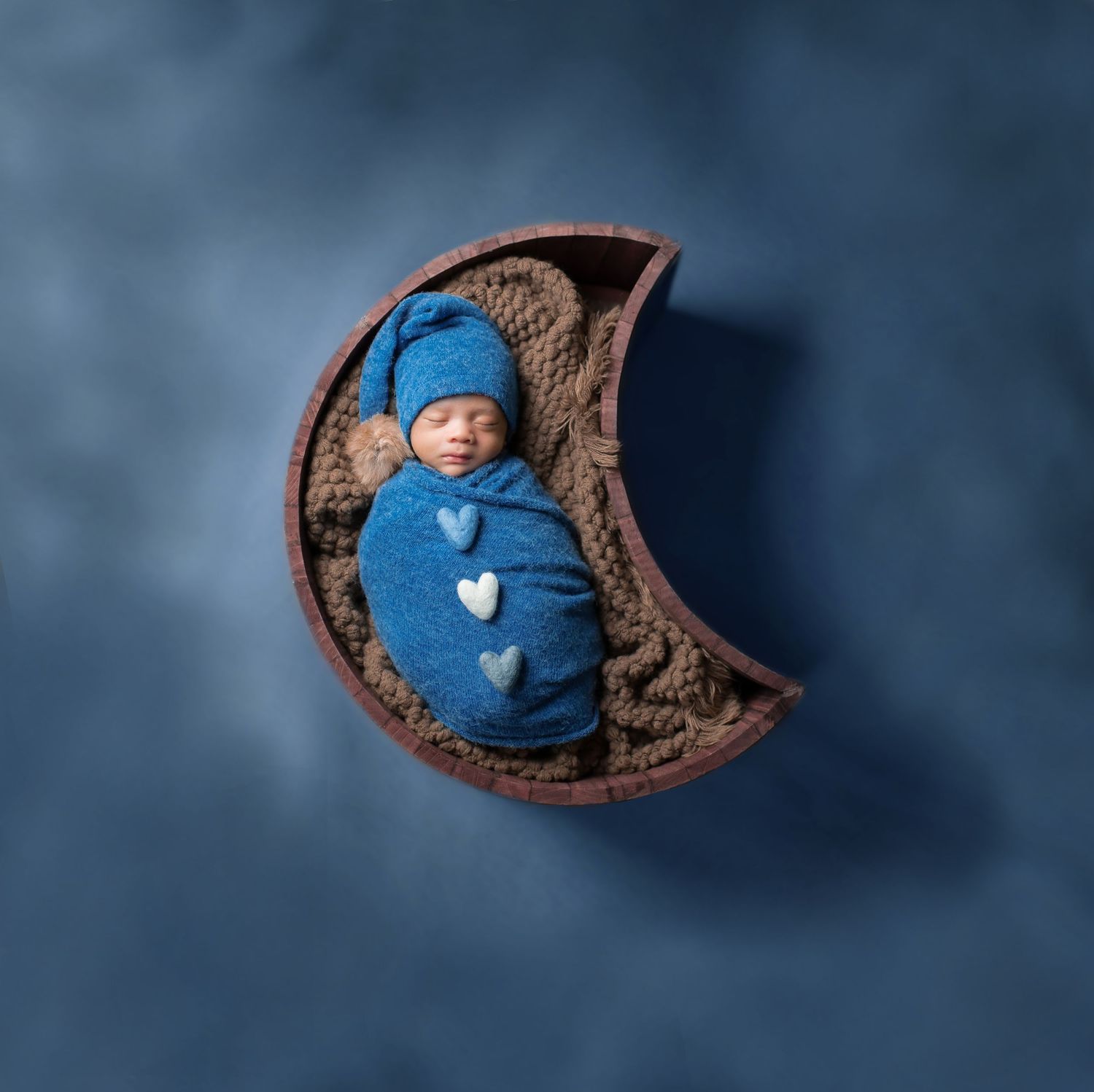 Newborn baby swaddled in blue wrap with heart details, sleeping in a crescent moon prop on a blue backdrop.