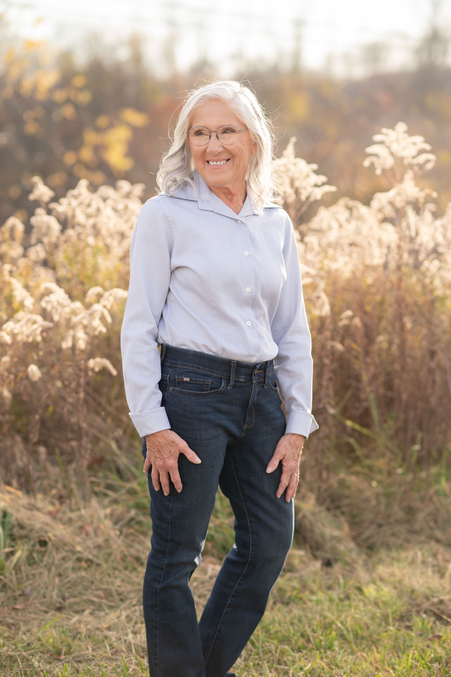 a lovely older woman in a blue shirt and jeans poses and smiles for a portrait in front of a field of golden sunlit grasses
