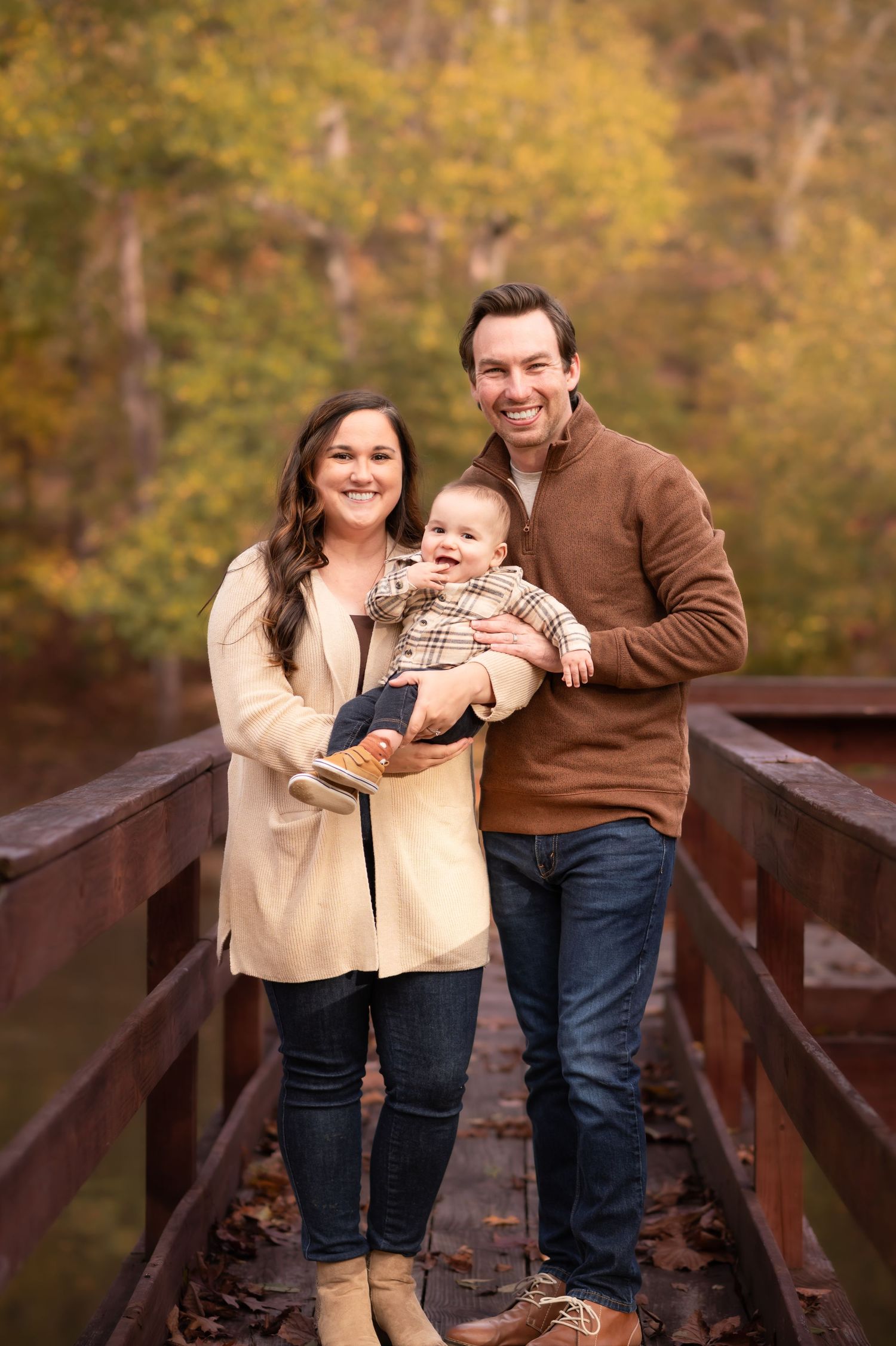 a young husband and wife and their baby boy pose together on a pier by a pond and smile for an autumn portrait, with fall leaves behind them