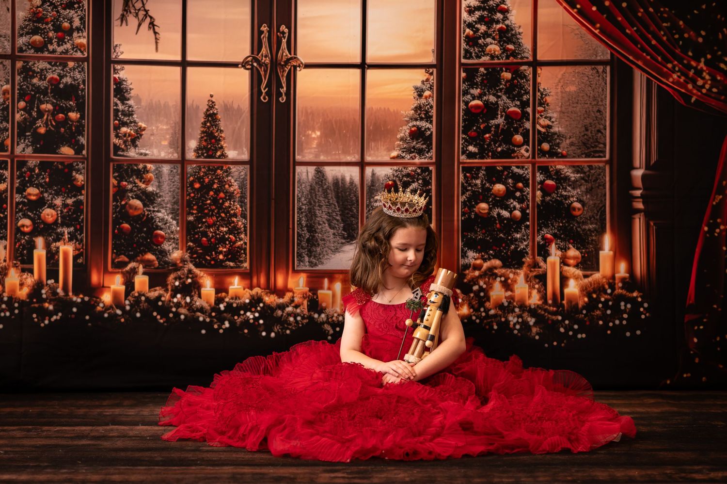 little girl with brown hair and a fancy red dress sits on a wood floor holding a nutcracker, with a candlelit window and snowy scene behind