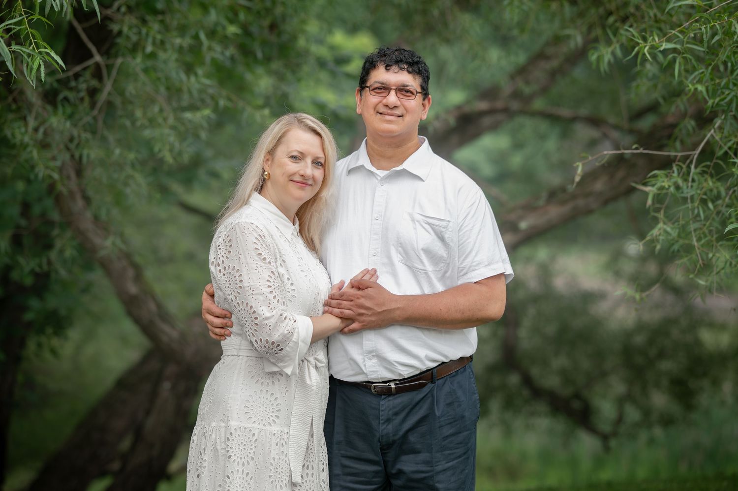 a husband and wife dressed in black and white pose together for a portrait, smiling and the camera and holding hands, with green background