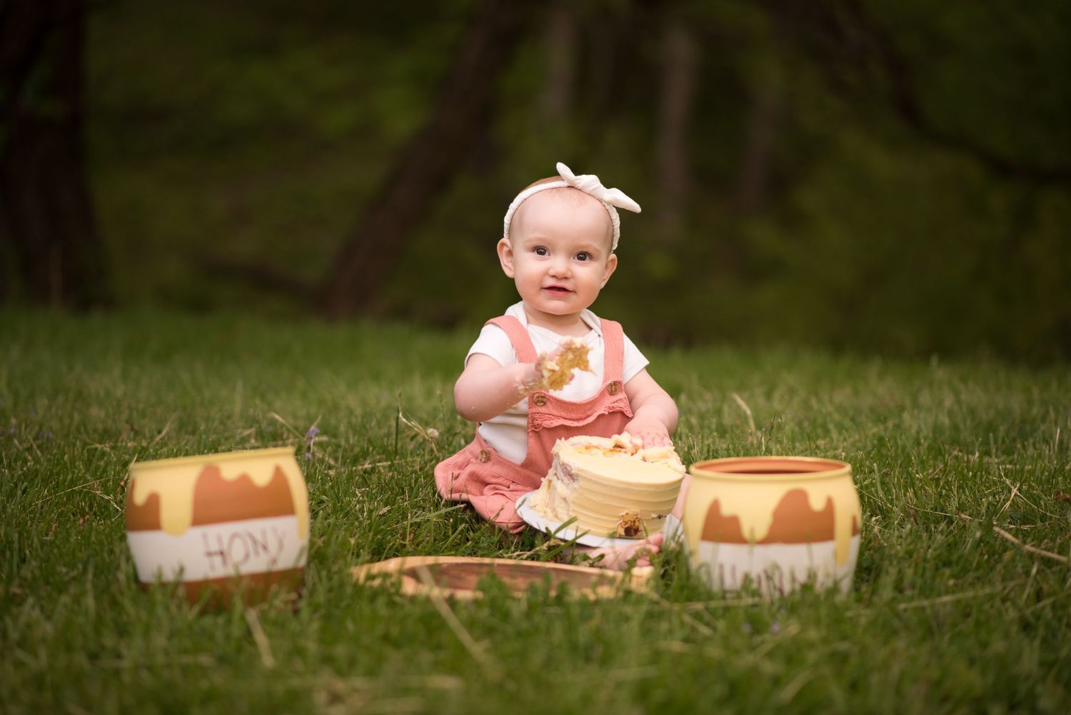 a cute toddler girl in orange overalls sits with two Winnie the Pooh honeypots in the grass and smiles for a birthday portrait