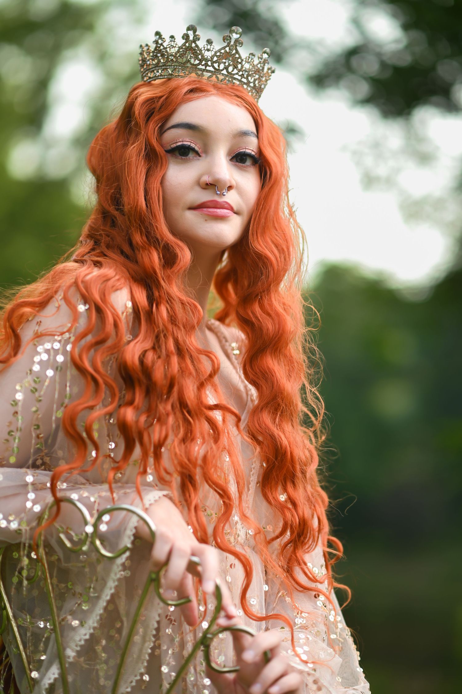 closeup portrait of a young woman with long red hair in a gold sequin gown and crown, looking down at the camera with a slight smile