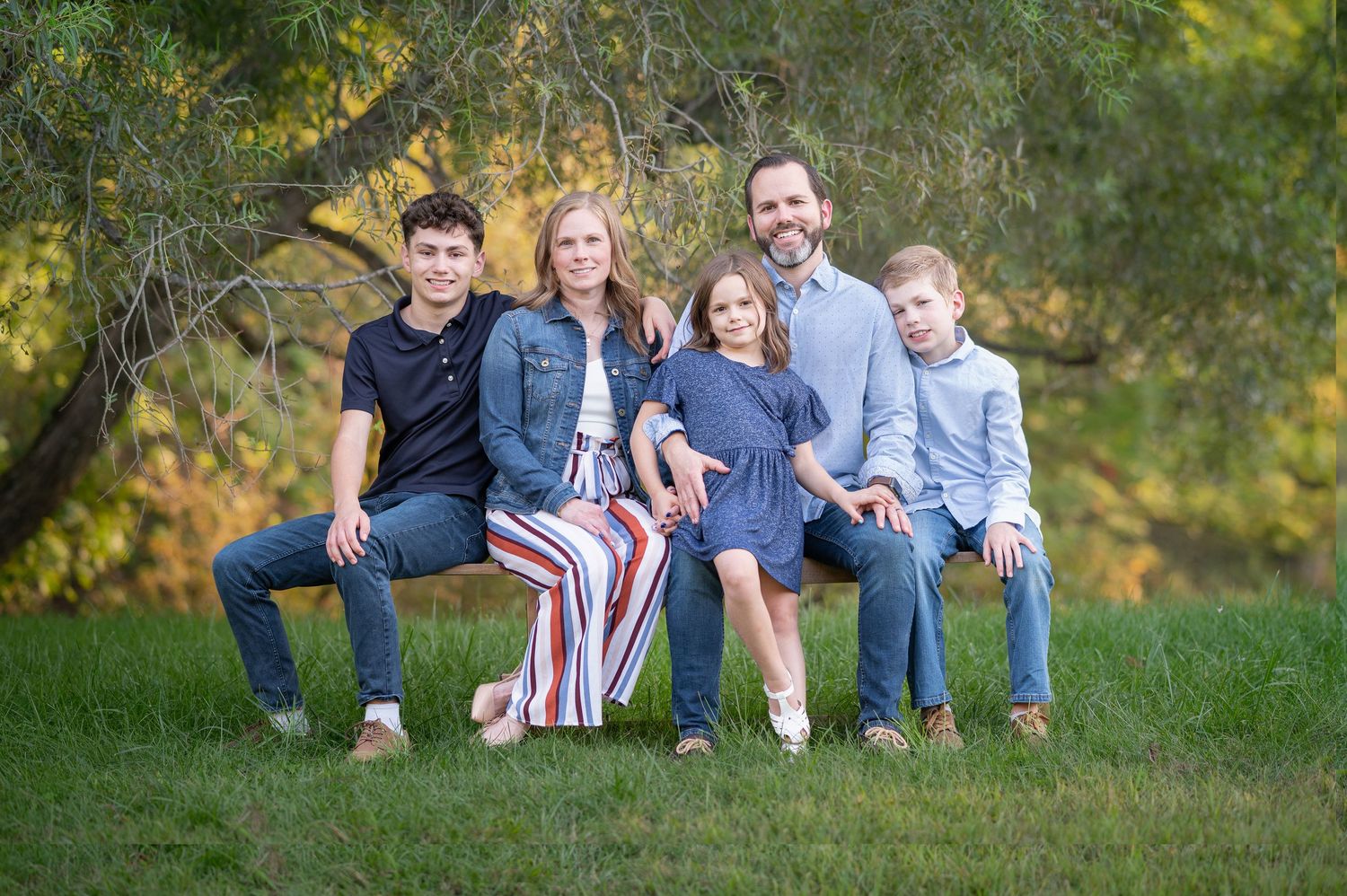 a family of five poses together on a wood bench in the grass under a large tree, with sunlit scenery in the background; Morgantown, WV