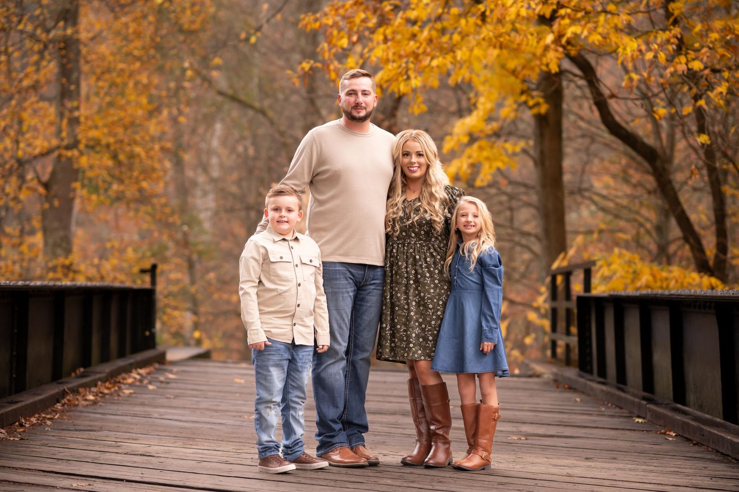 a family of four in jeans and cream sweaters poses for a portrait on an old wooden bridge with fall trees in the background; Fairmont, WV