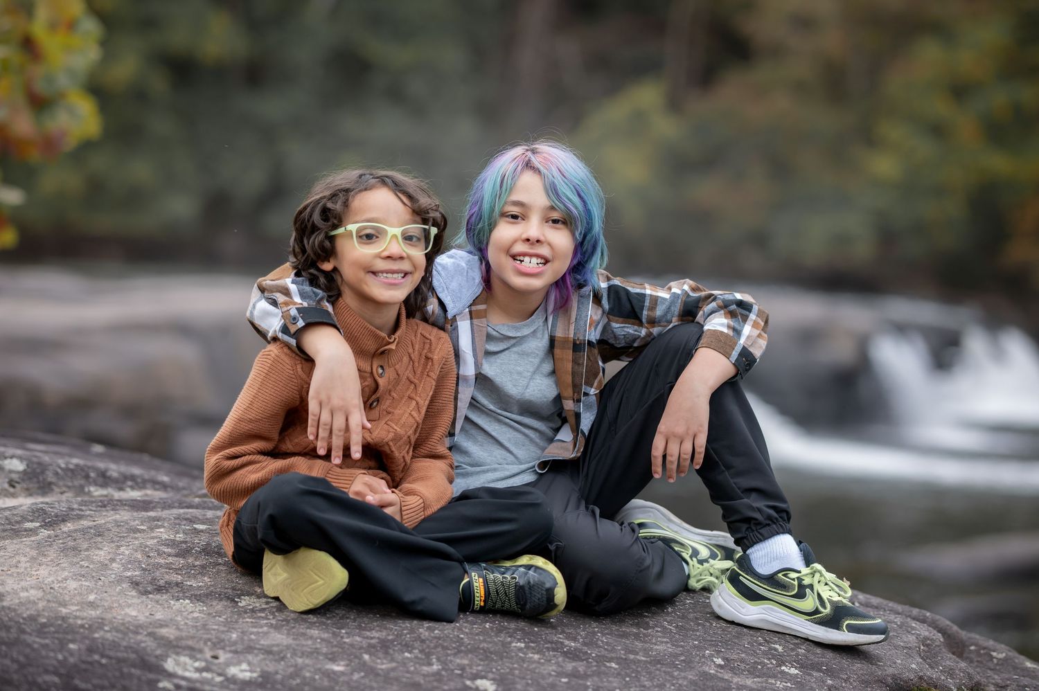 Two brothers smile with arms around each other for a portrait, sitting on rocks in front of waterfalls at Valley Falls State Park, WV