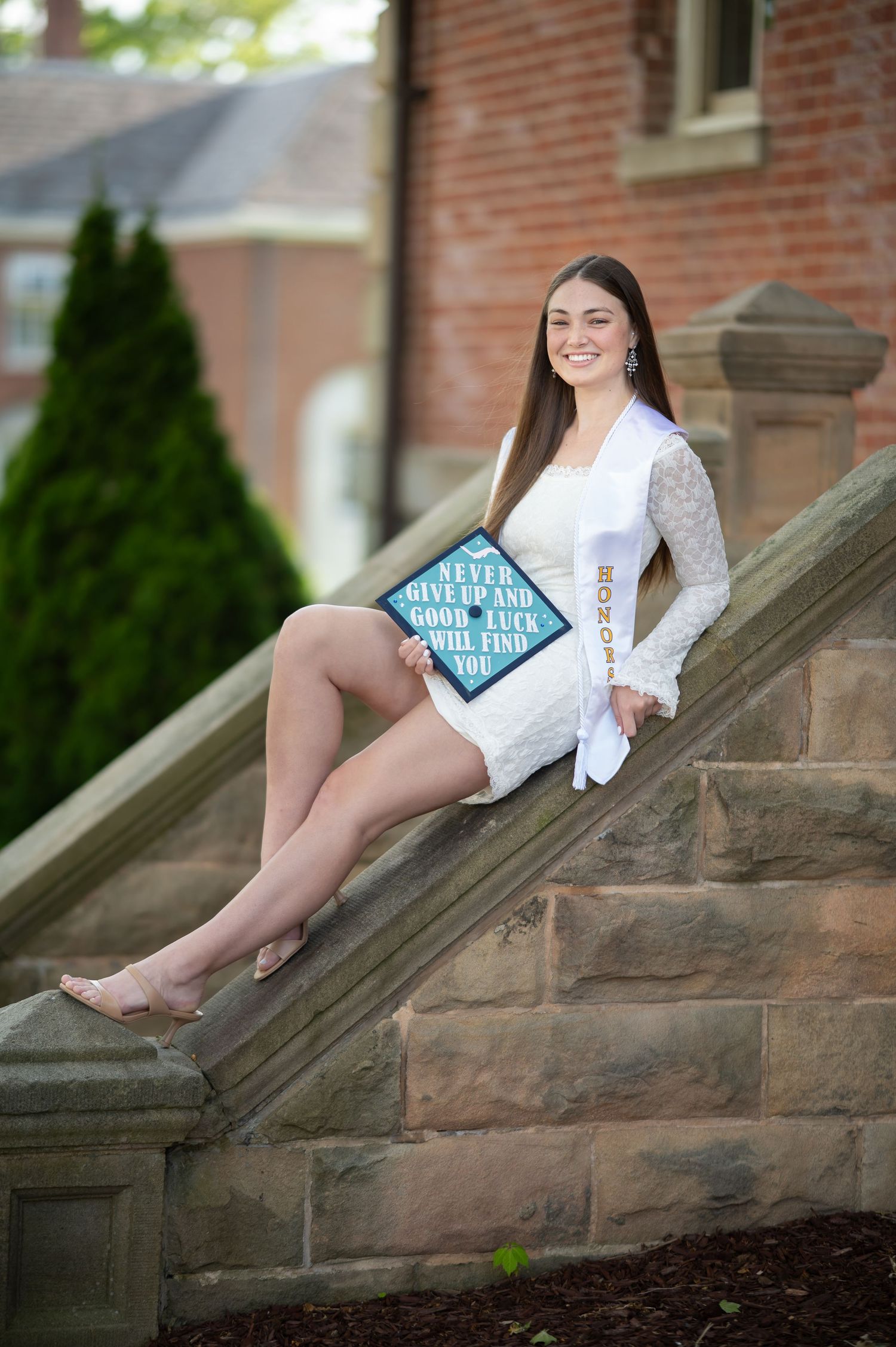 A WVU graduate student in a white lace dress poses with her decorated graduation cap on a set of stone stairs in Woodburn Circle