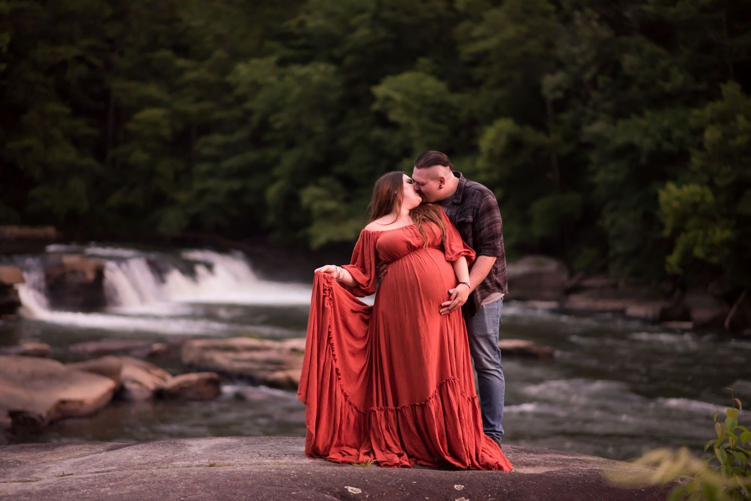 a pregnant woman in a long orange dress poses with her husband in front of waterfalls at Valley Falls State Park in Fairmont, WV