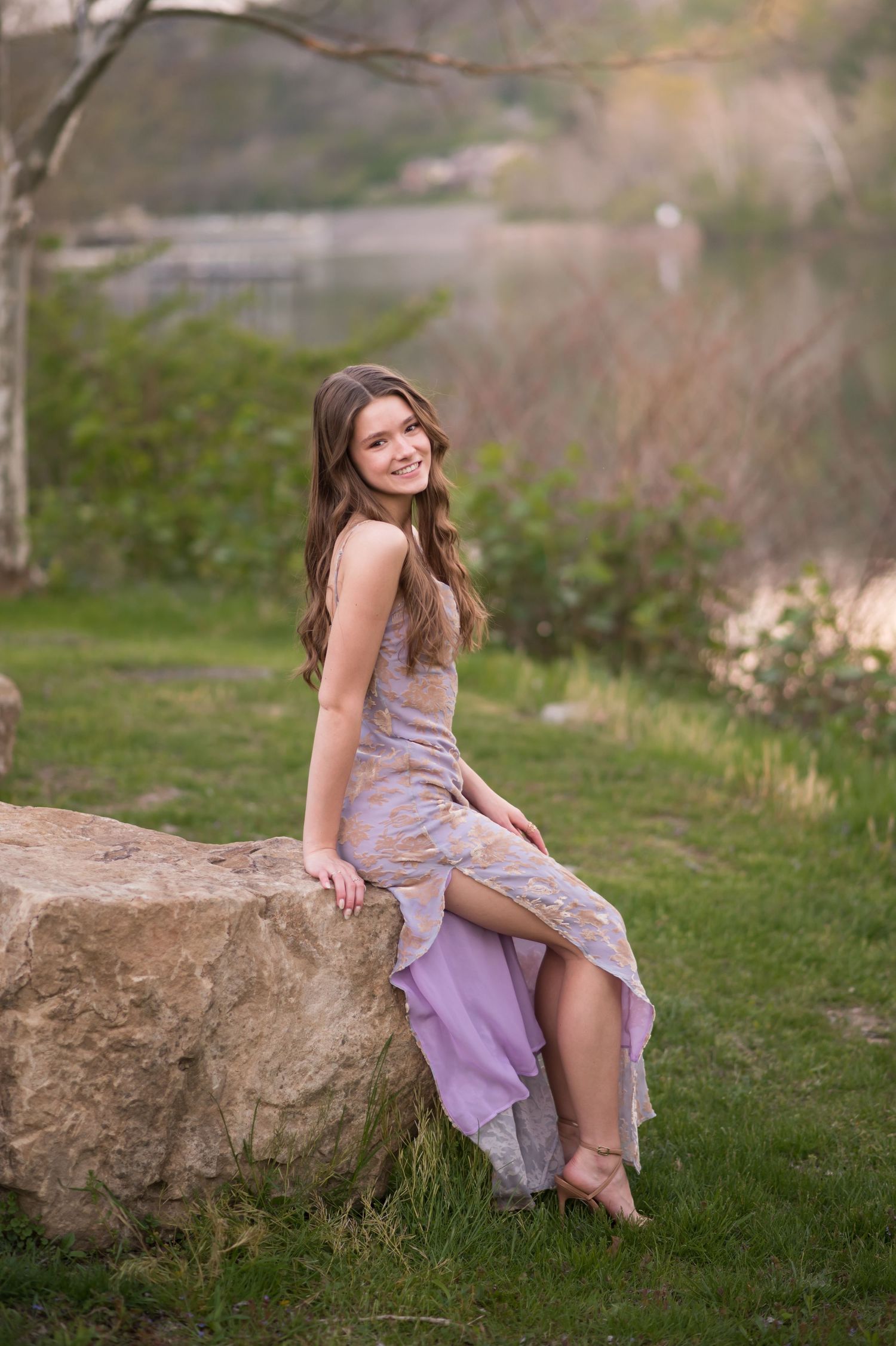 a young woman with dark hair and a long purple prom dress poses on a large rock in the grass in front of a river, with trees behind